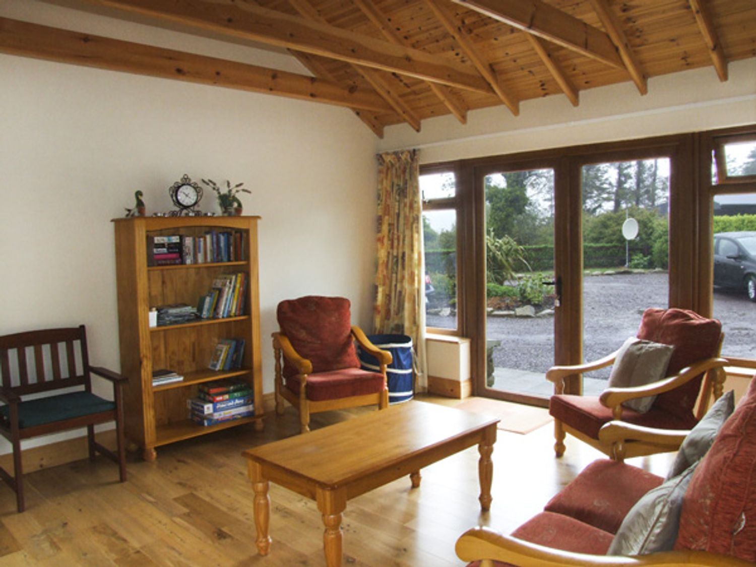 A living room with wooden ceiling and floor a bookshelf with books and a clock two armchairs and a coffee table near large glass doors at An Grianan in Clonakilty County Cork