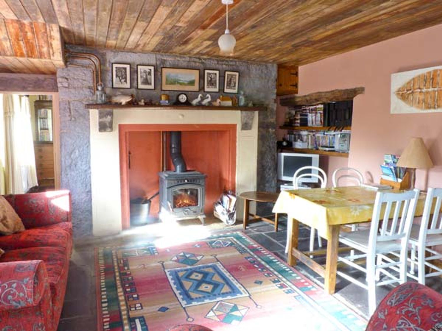 A living room with a wood burning stove a dining table with chairs a red patterned sofa and a colorful rug at Clooncorraun Cottage Ballinrobe County Mayo