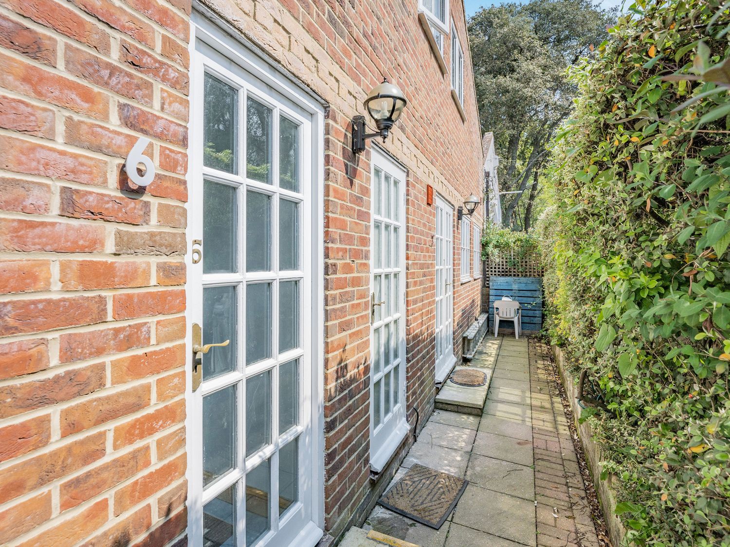 A brick exterior wall with white framed glass doors and outdoor lights along a narrow paved path with greenery on one side at Sail Loft Annexe in Yarmouth