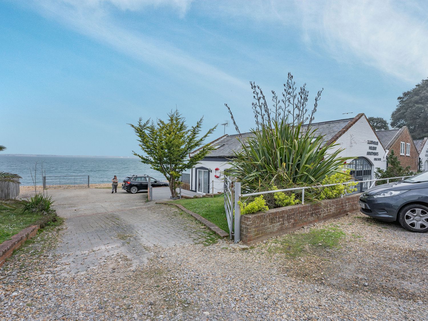 A gravel driveway leading to a parking area by the sea with parked cars and a white building with plants at Sail Loft Annexe in Yarmouth