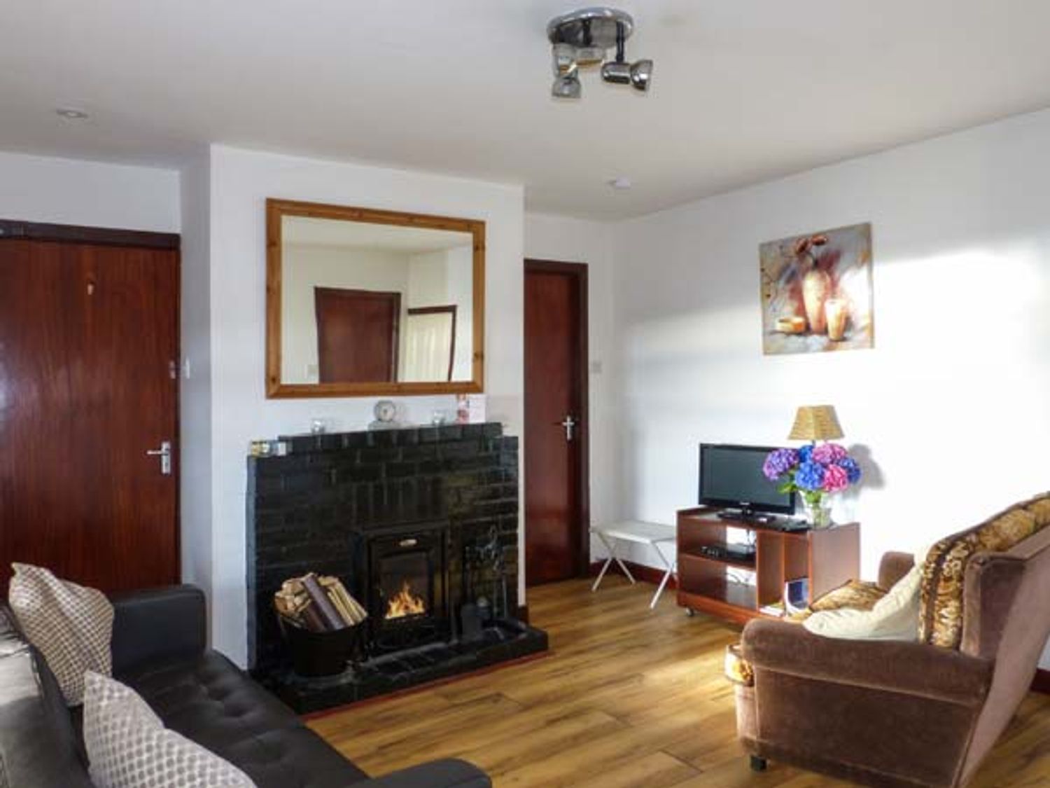 A room with a black fireplace a mirror above it a television on a wooden stand flowers on the stand and seating at Silver Strand Cottage Silver Strand near Wicklow Town County Wicklow