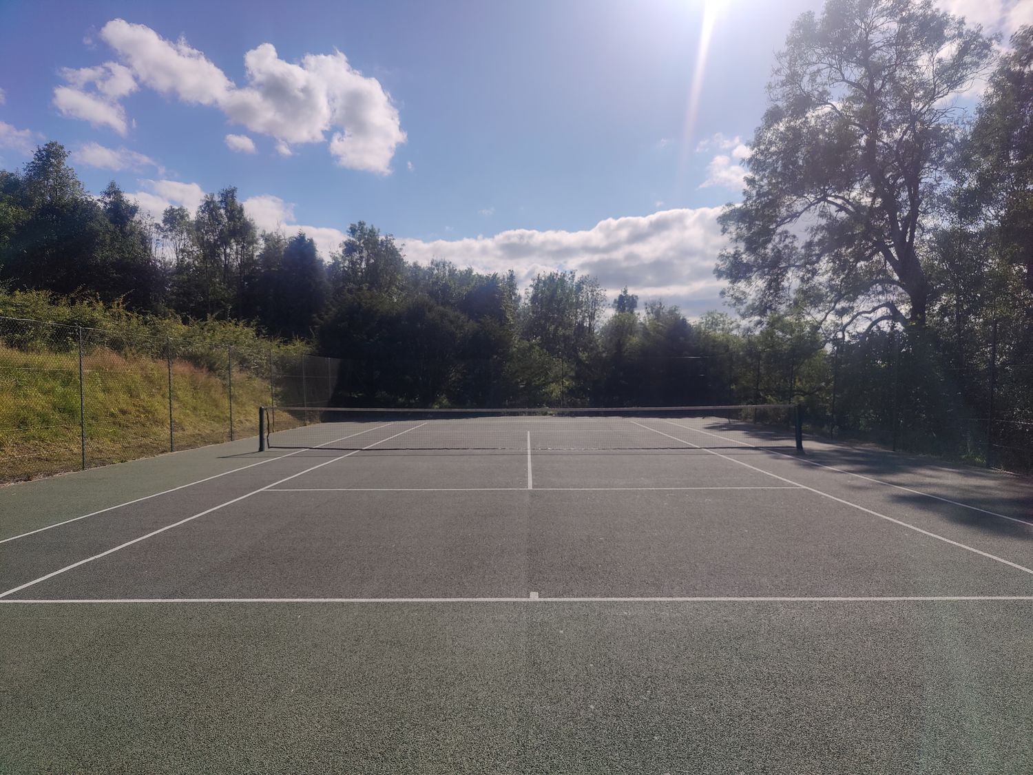 An empty outdoor tennis court surrounded by trees and a chain-link fence at Hiraeth in Dolau near Llandrindod Wells