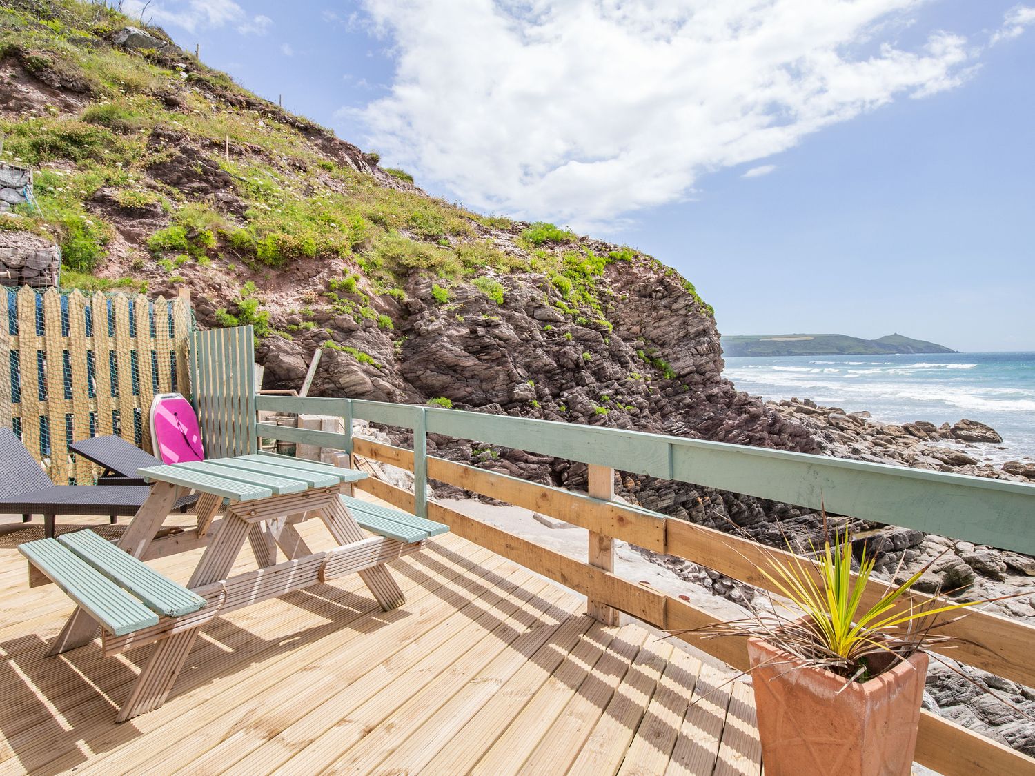 An outdoor wooden deck with a picnic table a plant a lounge chair and rocky cliffs by the ocean at Beachcomber's Cottage in Waberthwaite