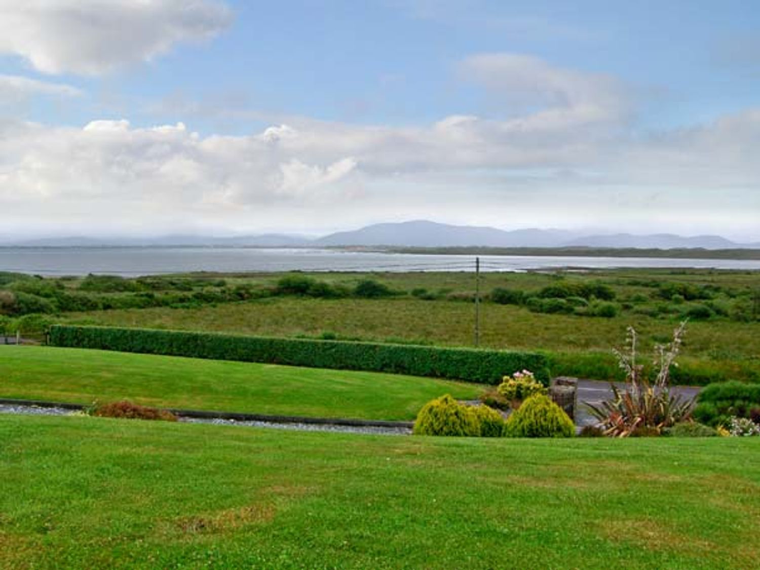 A grassy yard with bushes and a hedge overlooking a body of water and distant hills at Heron's Nest in Inch County Kerry