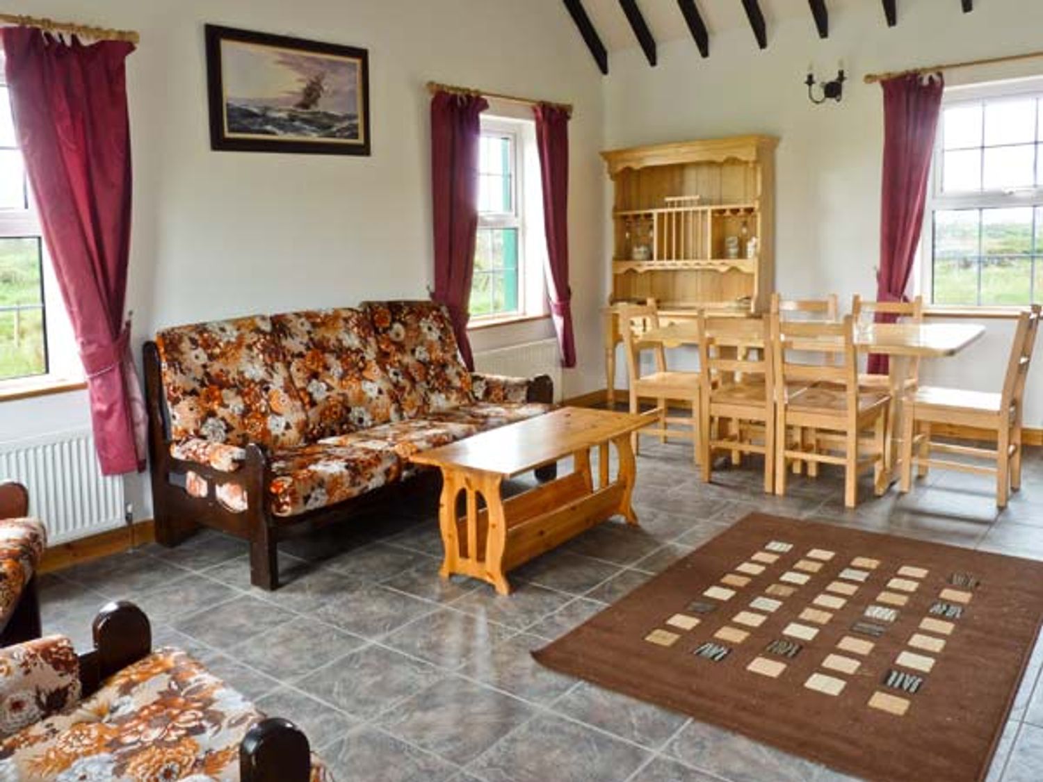 A living room with a floral patterned sofa a wooden coffee table wooden dining furniture and red curtains at Lully More Cottage in Cruit Island County Donegal