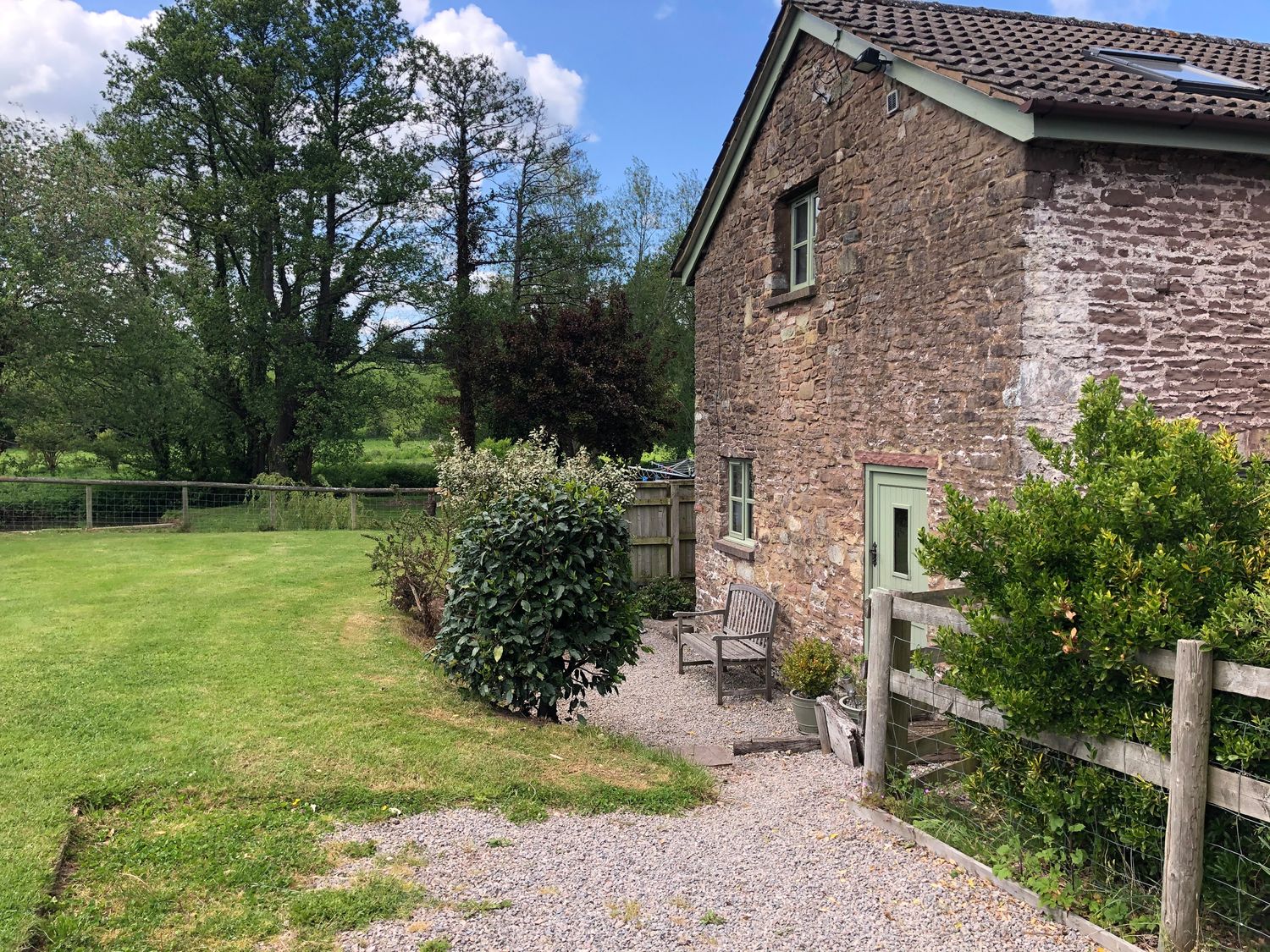 A stone house with green doors and windows next to a garden with trees and bushes at The Stable in Marstow near Ross-on-Wye