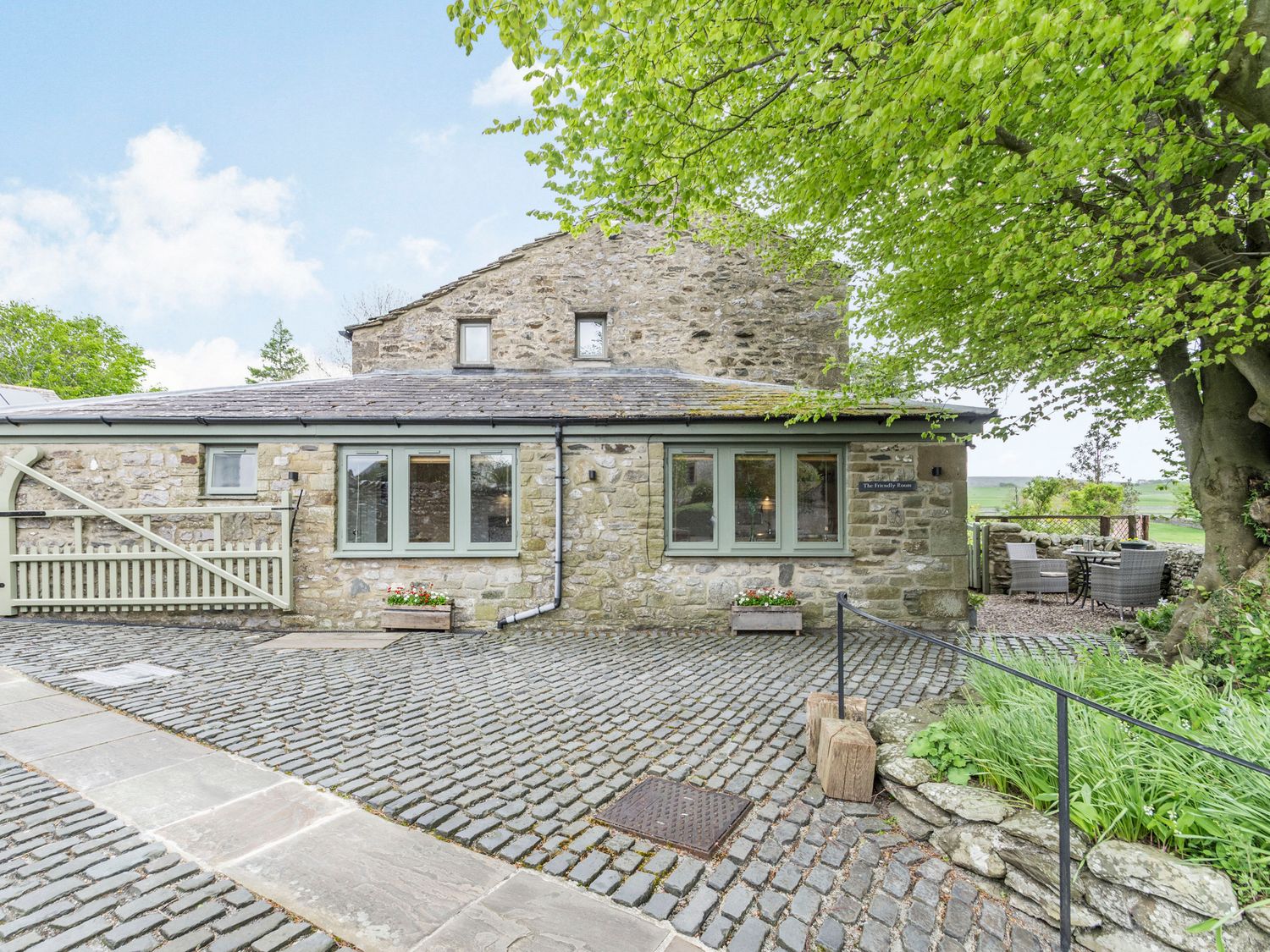 A stone cottage with green framed windows and outdoor seating under a tree at The Friendly Room in Austwick