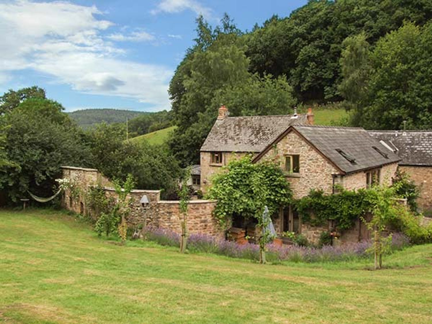 A stone house with a garden wall surrounded by grass and trees at The Lodge Farm Barn in Deepdean near Ross-on-Wye