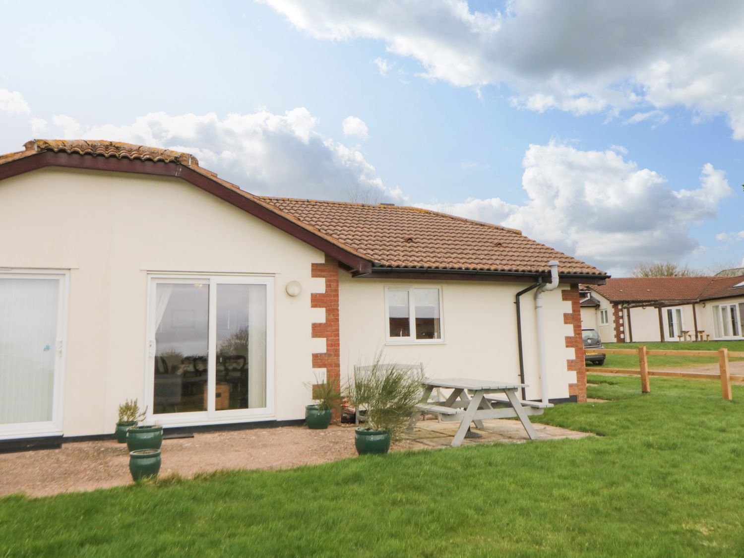 A single story house with a tiled roof green potted plants a picnic table and grass at Broom Cottage in Stoneleigh Holiday Village near Sidmouth