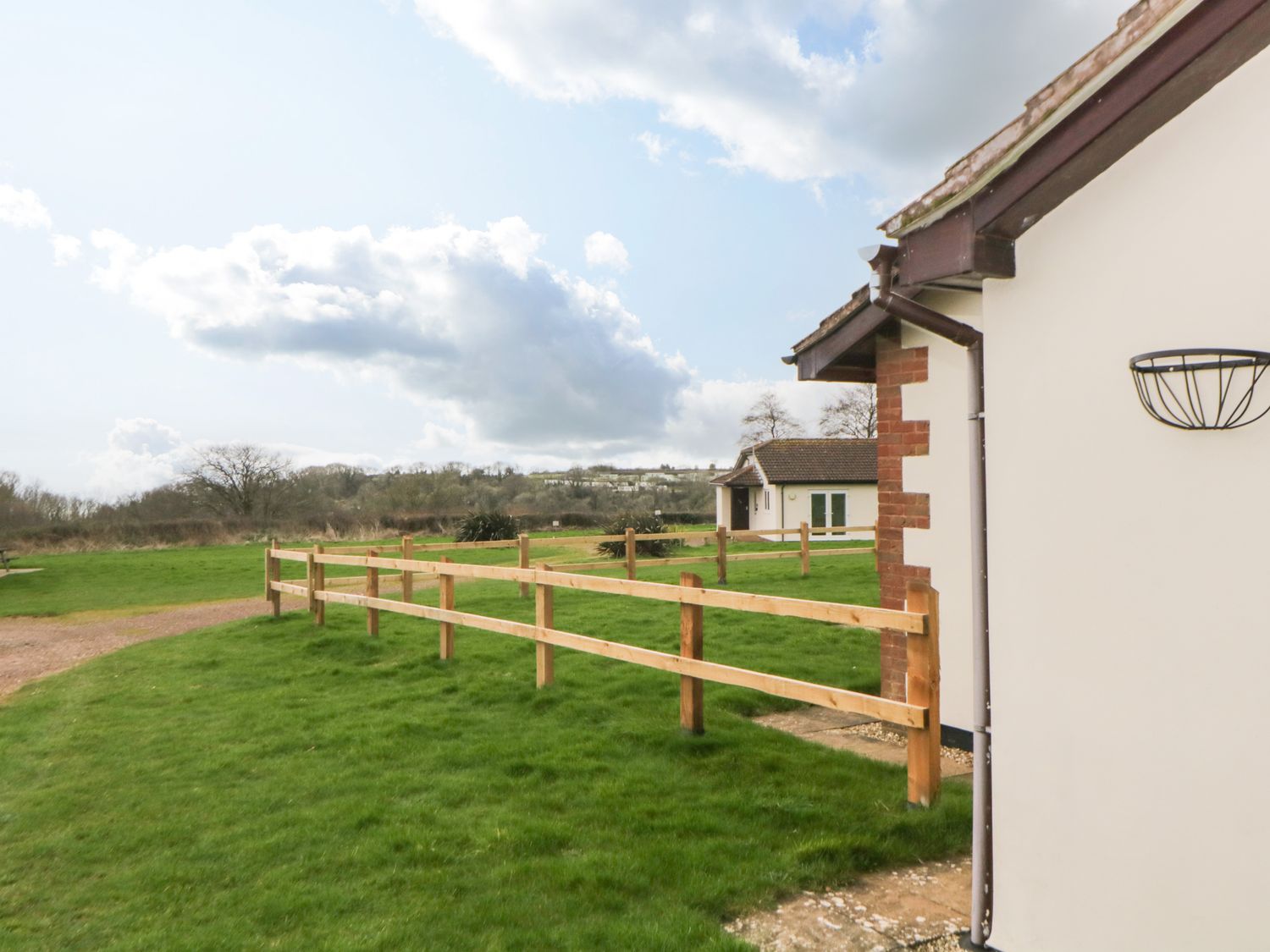 View of a grassy outdoor area with wooden fences and white buildings under a cloudy sky at Broom Cottage in Stoneleigh Holiday Village near Sidmouth