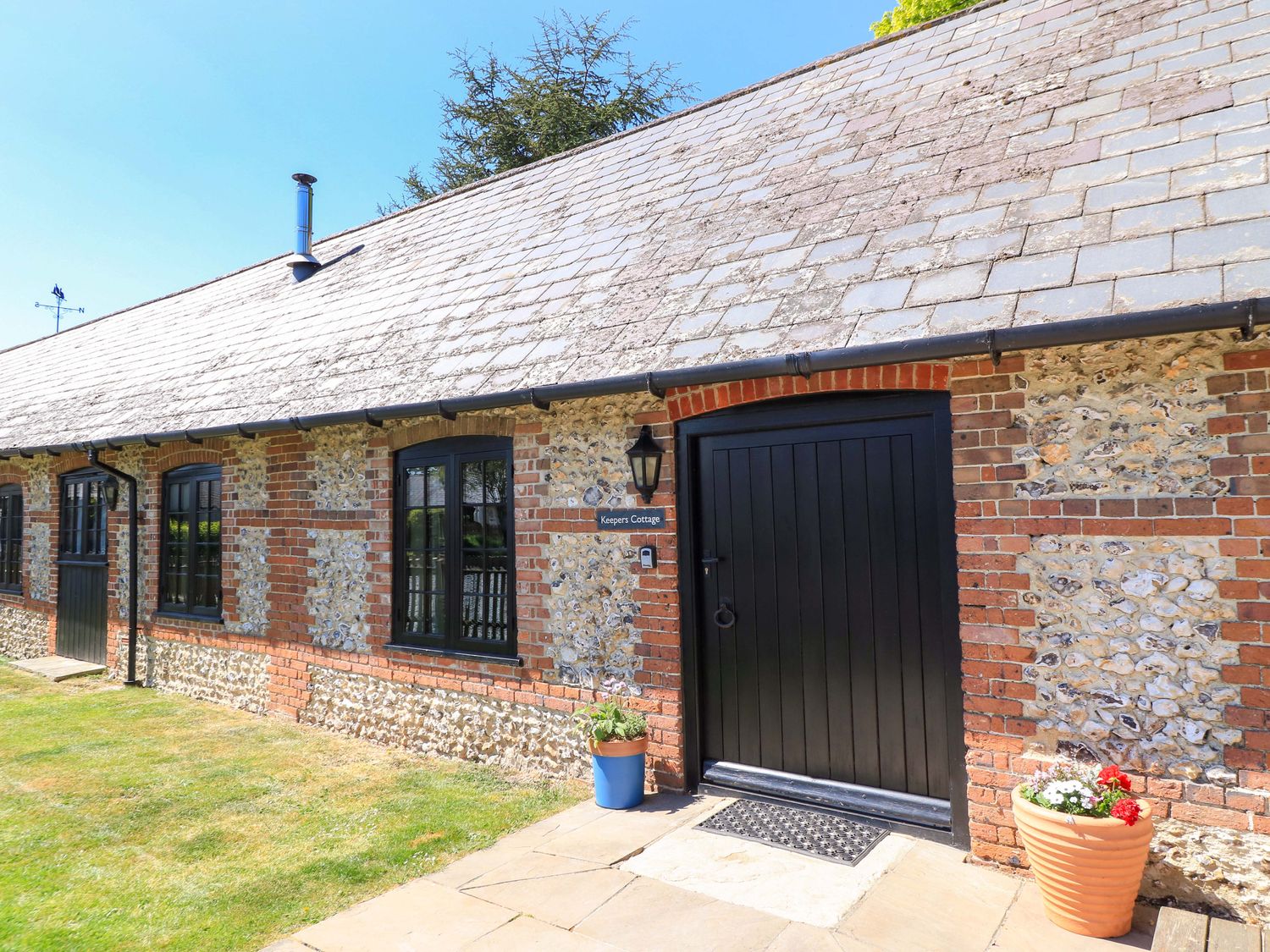 Exterior of a single story stone and brick cottage with black windows and doors and two flower pots at Keepers Cottage in Winterborne Stickland