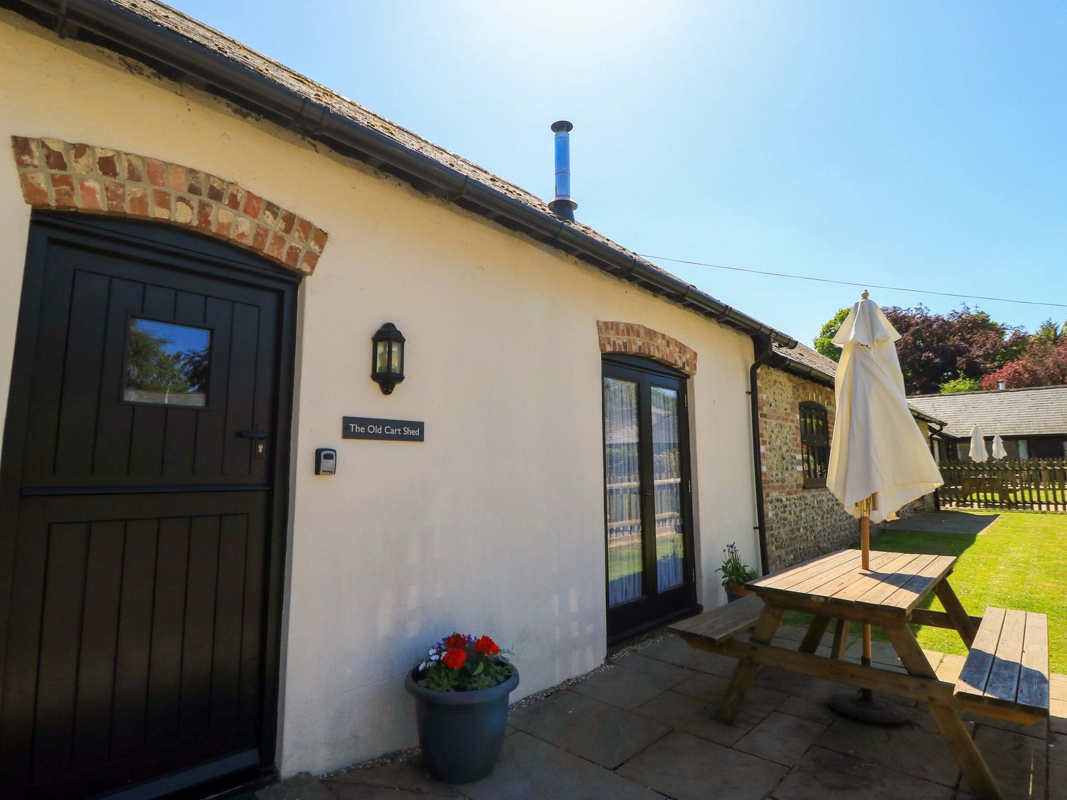 A patio area with a wooden picnic table and umbrella outside a building with black doors at The Old Cart Shed in Winterborne Stickland