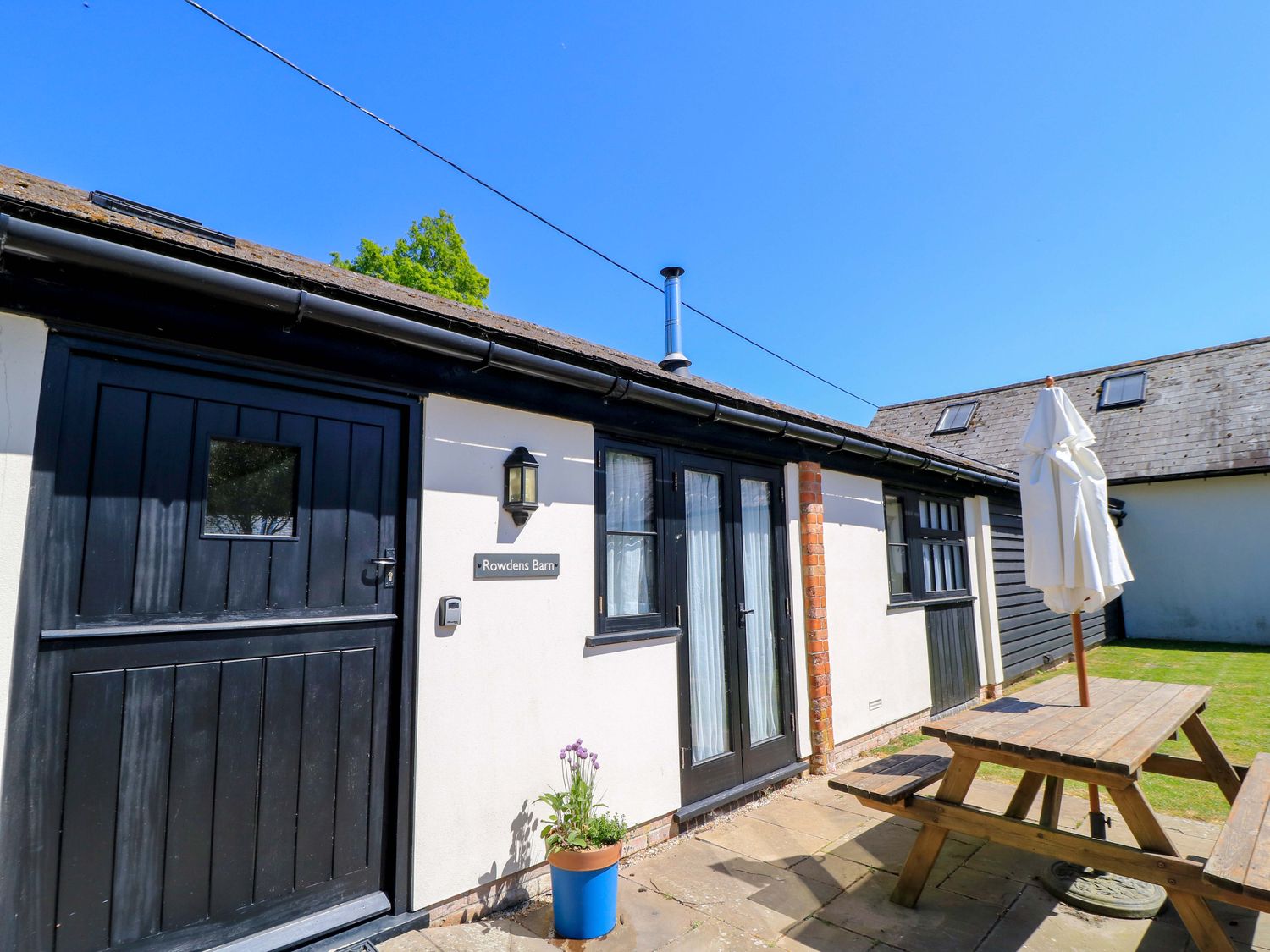 Exterior view of a building with black doors and windows a wooden picnic table and umbrella on a patio at Rowdens Barn in Winterborne Stickland