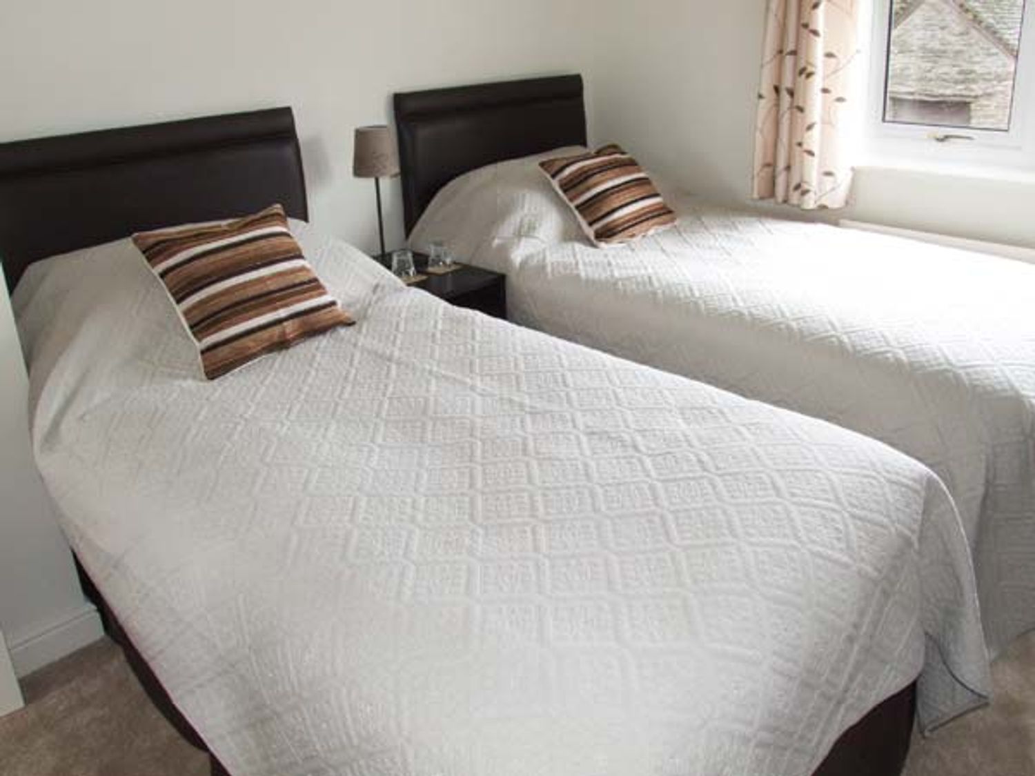 A bedroom with two single beds with white bedspreads and striped pillows a bedside table with lamp and glasses and a window with curtains at Hour Cottage in Stow-on-the-Wold