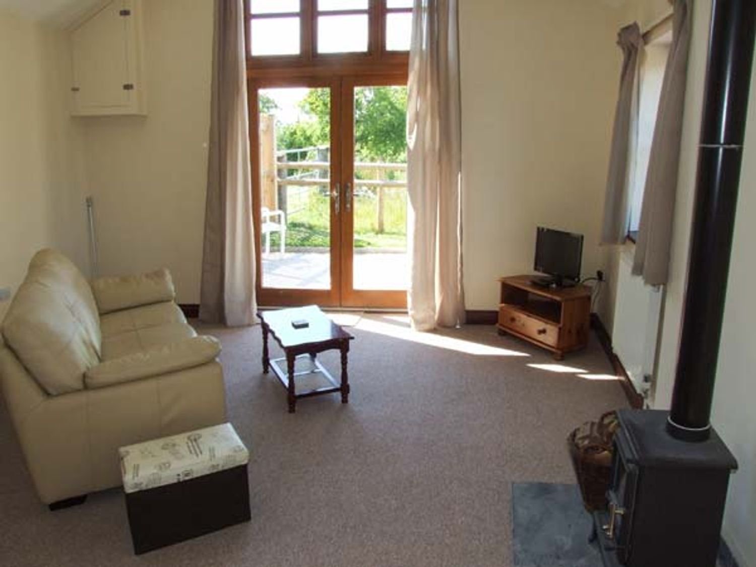 A living room with a beige sofa small wooden coffee table television on a wooden stand wood burning stove and glass doors at Dovetail in Westbury-on-Severn