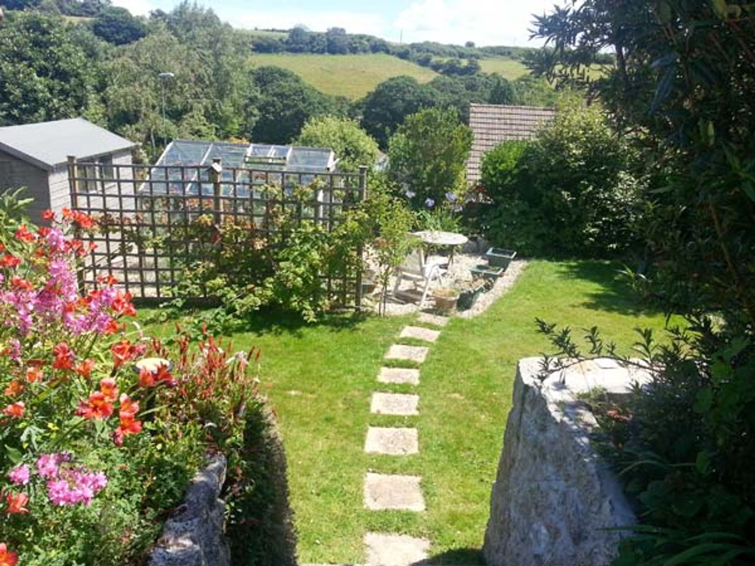 A garden area with a stone path leading to a seating area with a table and chairs surrounded by plants and flowers at Joylands in Falmouth
