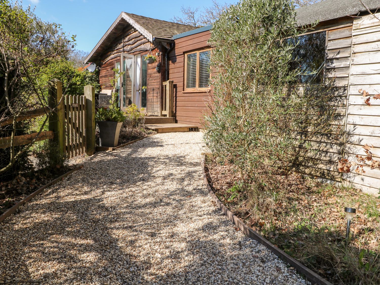 A gravel pathway leading to a wooden house with plants and a wooden fence at Woodmancote Lodge in Linchmere near Haslemere