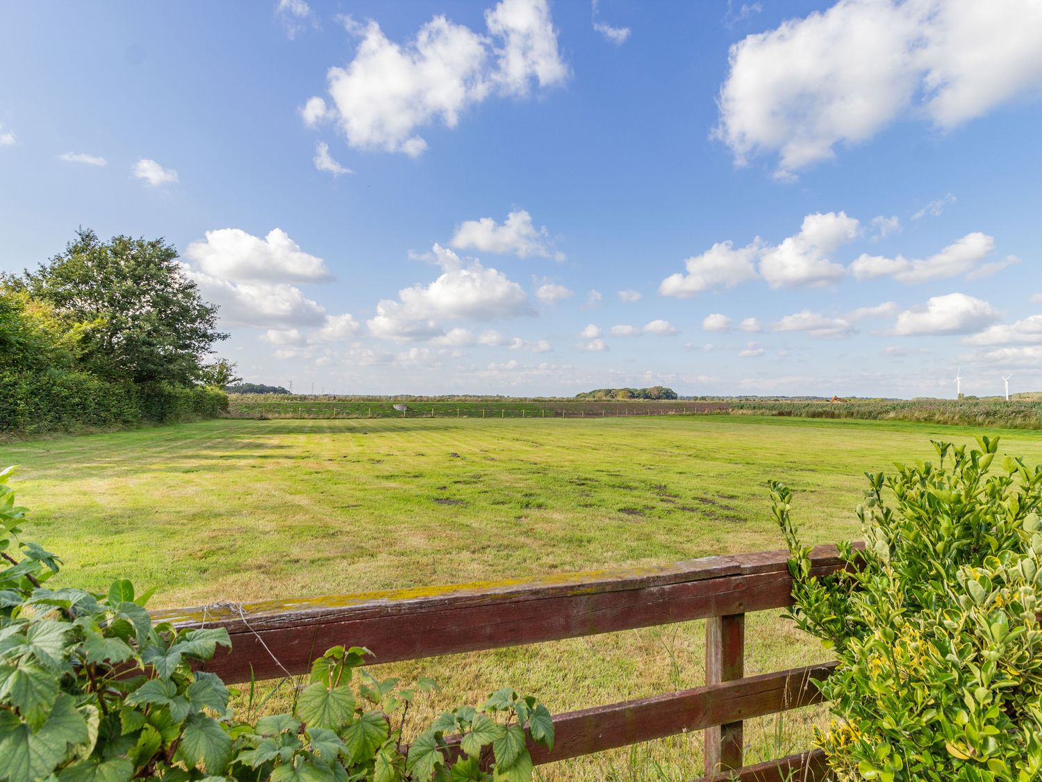 A fenced grassy field with bushes and trees under a blue sky with clouds at Wild Duck Lodge in Mawdesley near Southport
