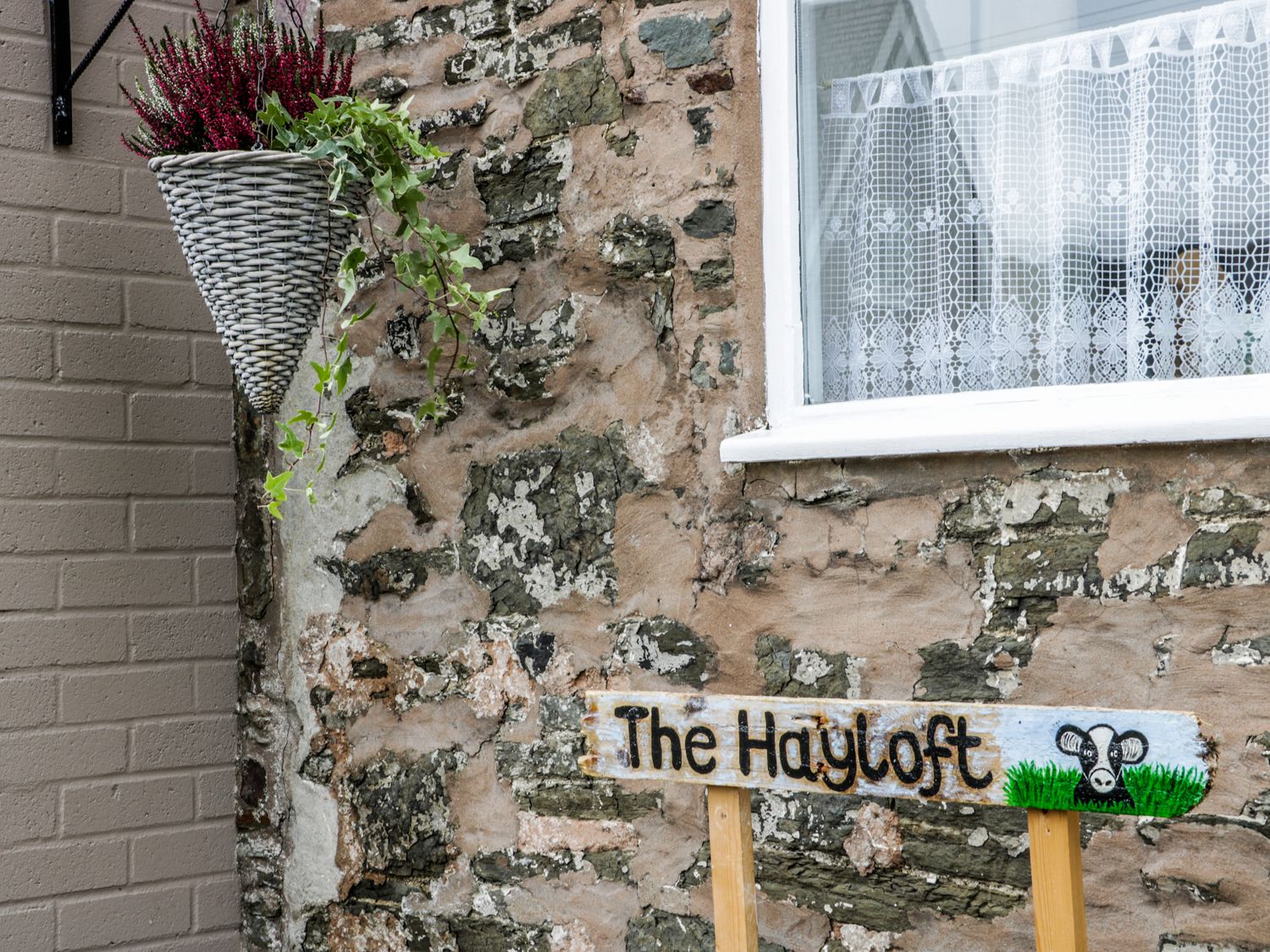 A stone wall with a lace curtain window a hanging basket with plants and a wooden sign that says The Hayloft in Stiperstones
