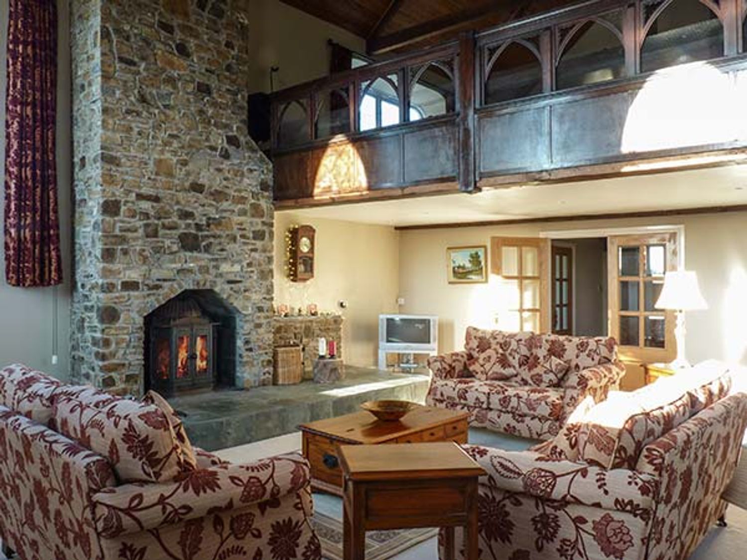 A living room with a stone fireplace sofas with floral covers wooden tables a TV and a balcony at Old Chapel in Clovelly