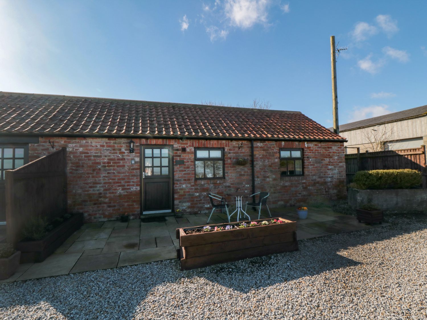 A brick exterior of a single-story building with a tiled roof a wooden door two windows and a small patio with two chairs and a table at Blakey in Gerrick near Moorsholm