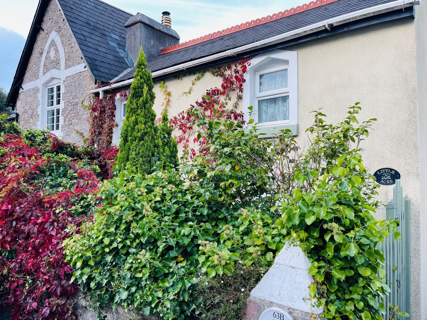 The exterior of a house with stone and cream walls surrounded by green and red foliage at Little Acre in Torquay
