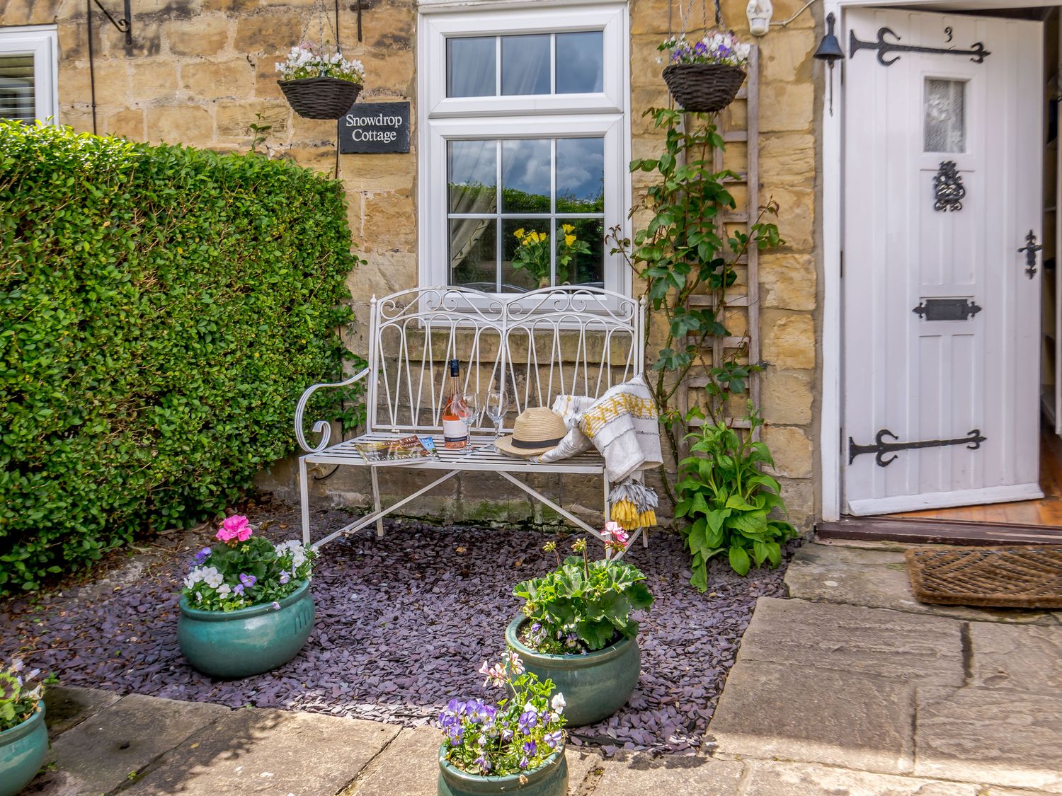 A white bench with a hat, bottle, glass and cloth outside a stone cottage with potted flowers at Snowdrop Cottage in Clifford West Yorkshire