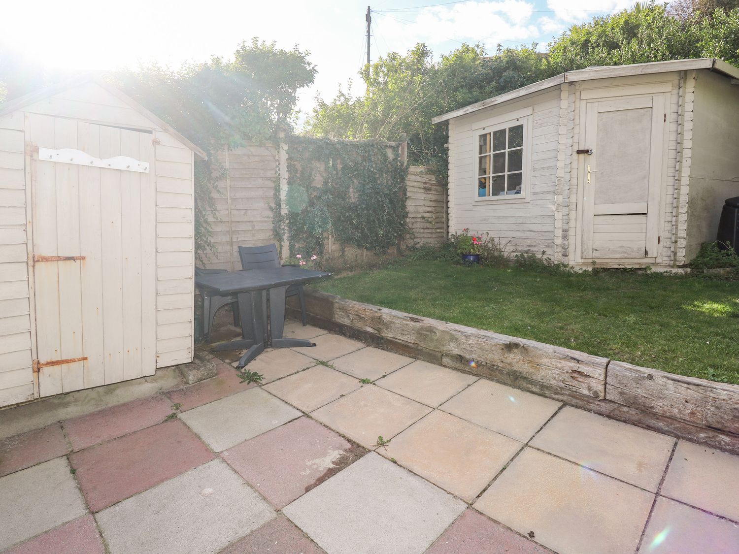 A garden with a white shed grass area wooden fence plastic table and chair on a tiled patio at Driftwood Polzeath near Wadebridge