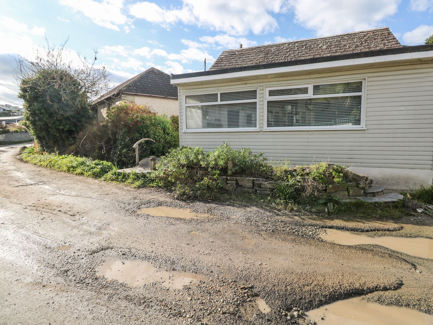 A gravel road with puddles beside a building with horizontal siding and large windows at Driftwood in Polzeath near Wadebridge