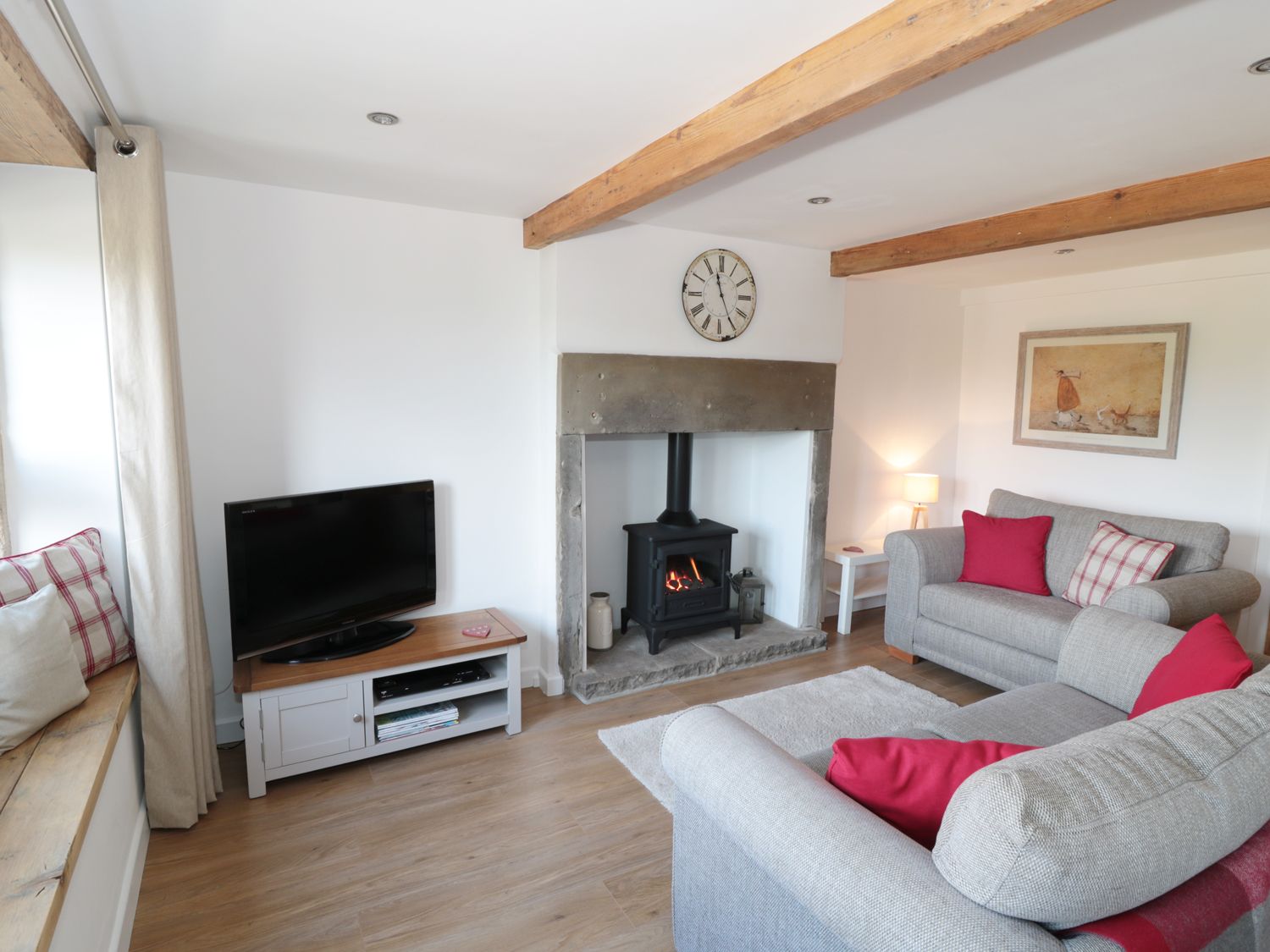 A living room with a wood burning stove TV on a stand sofas with red cushions and wooden beams on the ceiling at Bramble Cottage in Holmfirth