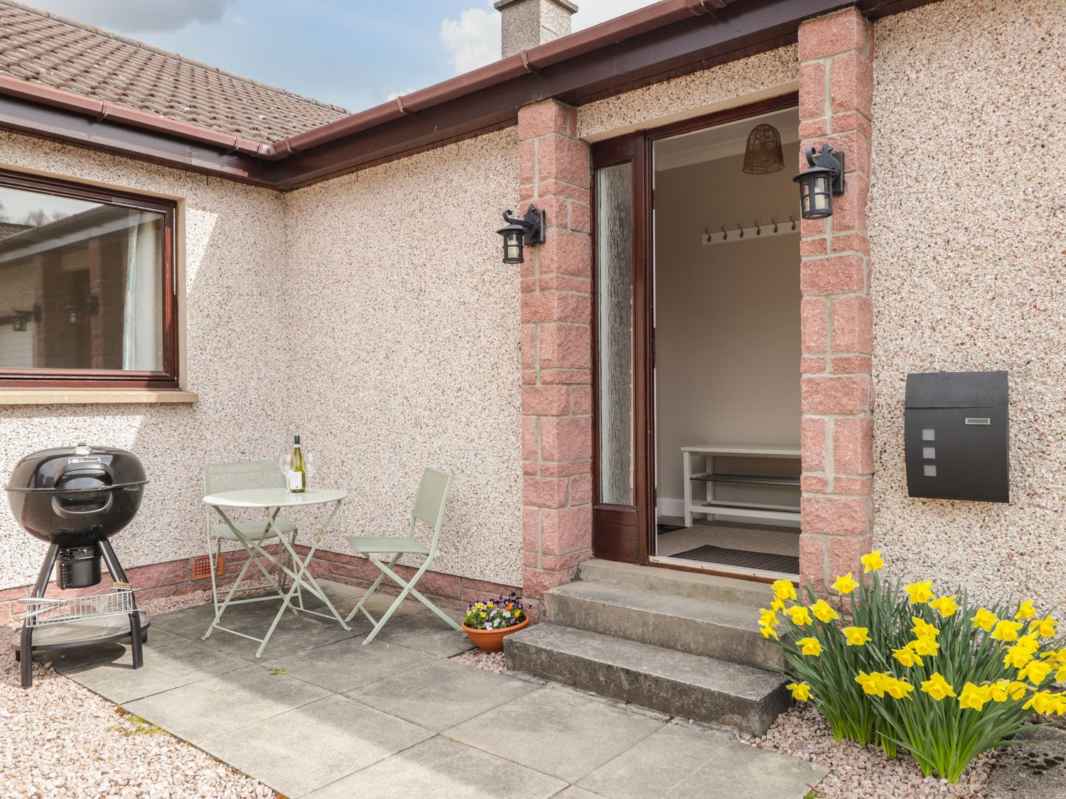 An outdoor patio area with a small table and two chairs a black barbecue grill and yellow flowers near the steps to a house entrance at Millview in Boat of Garten