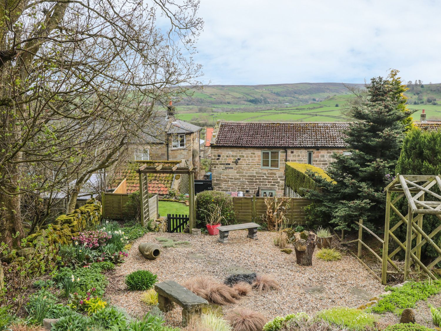 A garden with gravel ground benches plants and stone walls with houses and hills in the background at New Stable Cottage in Lealholm