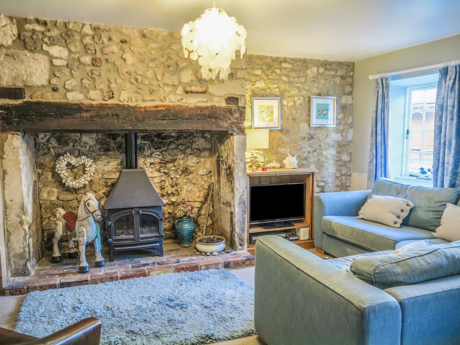 A living room with stone walls a wood stove a rocking horse and blue sofas at Craft Cottage in Beer near Seaton
