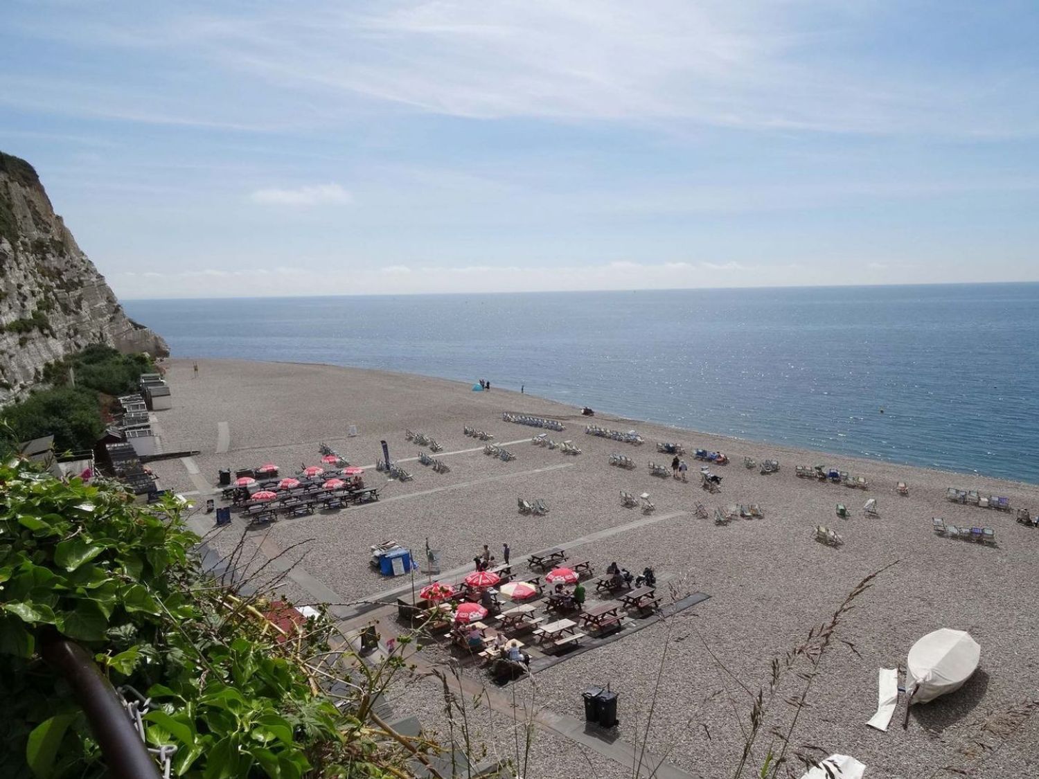A pebble beach with sun loungers tables with red umbrellas and a covered boat at Craft Cottage Beer near Seaton