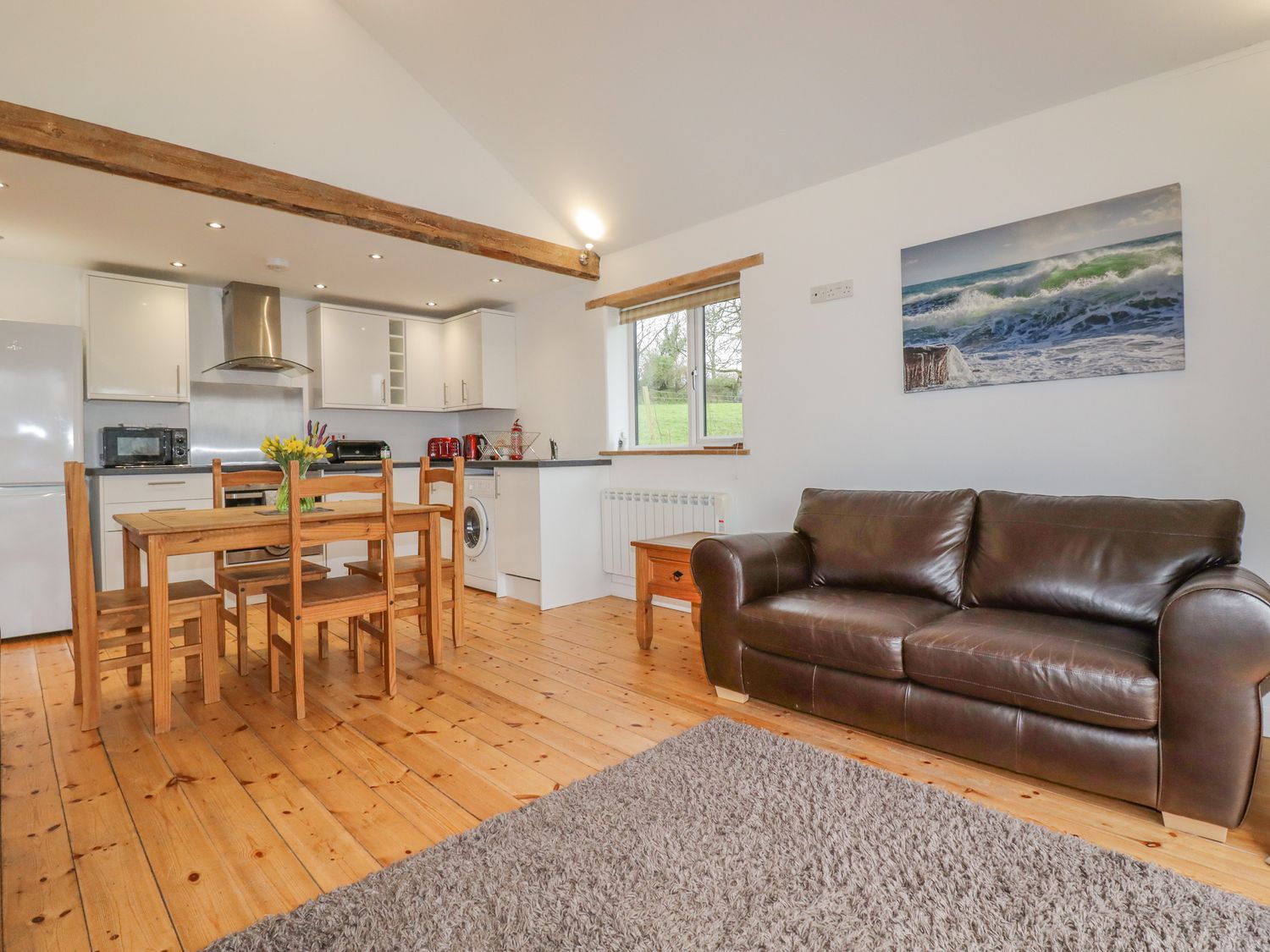 An open plan kitchen and living area with wooden dining table and chairs a brown leather sofa and kitchen appliances at Vine Cottage in St Ewe near Sticker 