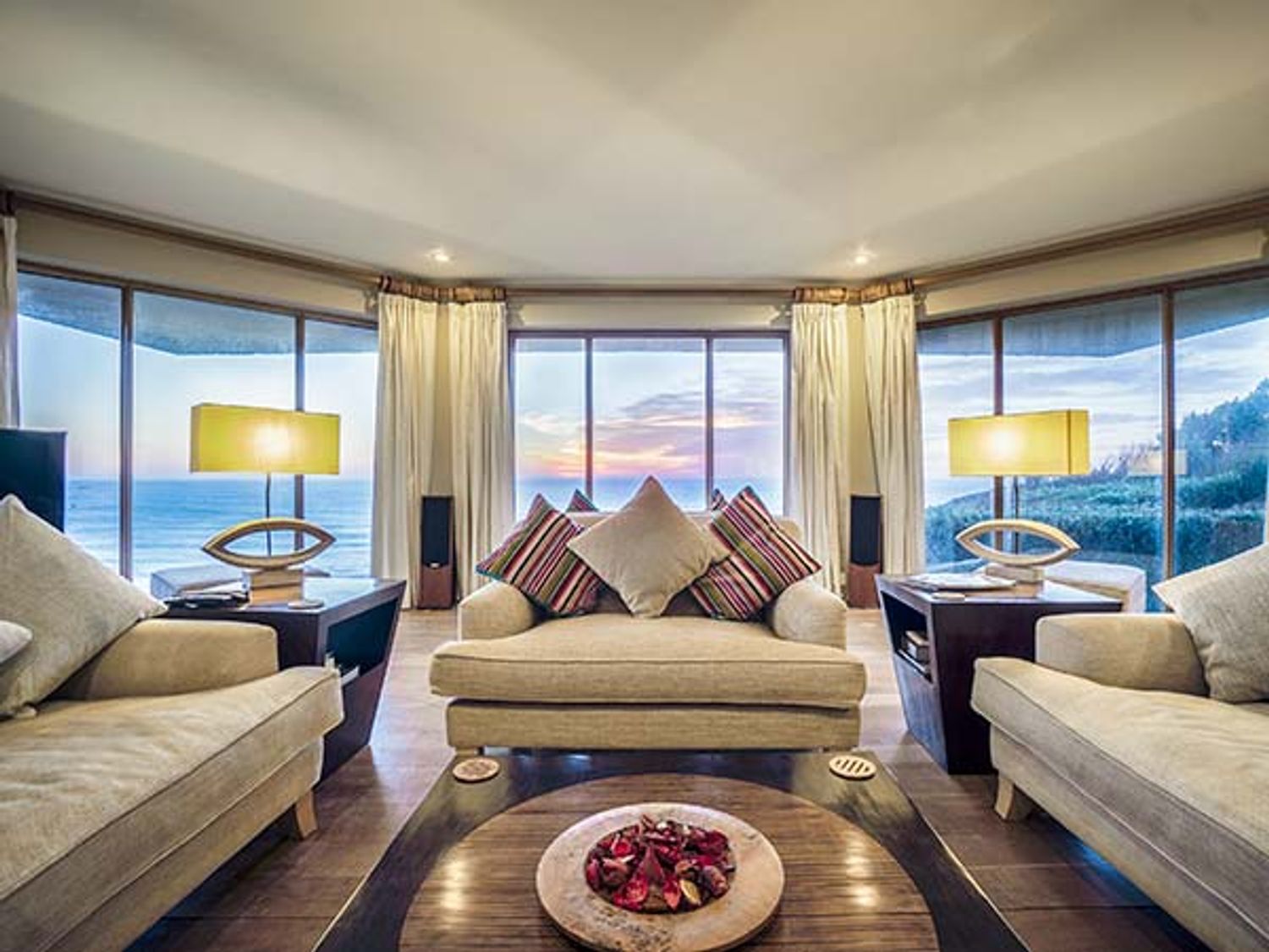 A living room with beige sofas and striped cushions overlooking the ocean through large windows at Cragford in Sennen Cove