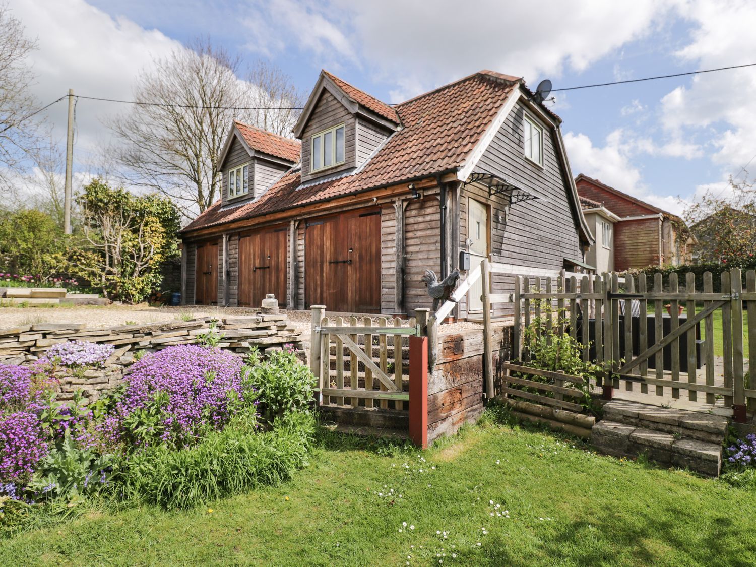 A wooden house with three garage doors surrounded by a garden with purple flowers and green grass at Shephard's Retreat in Charlton Mackrell near Somerton