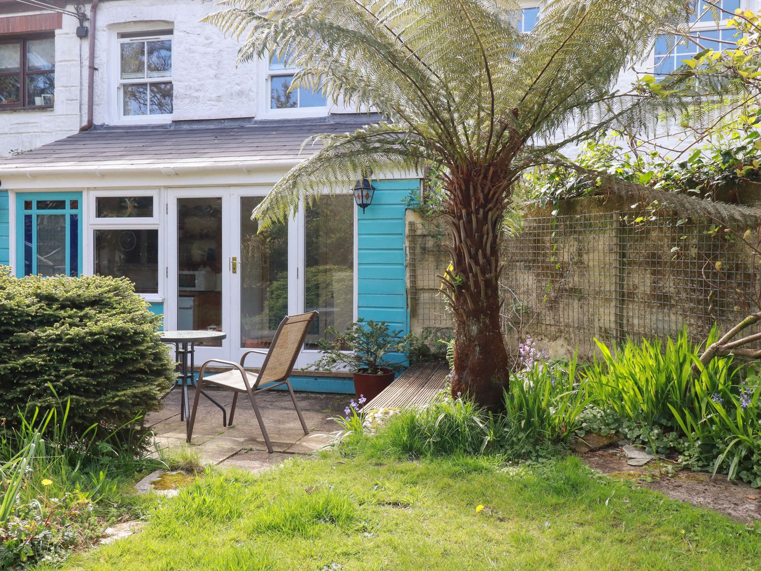 A garden with a tree a bush and a chair and table set outside a house with glass doors at The Ark Cottage in St Blazey