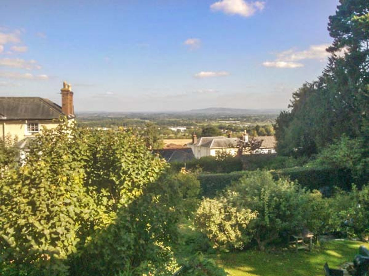 A garden with trees and shrubs and houses in the distance with a view of the countryside at Woodland Cottage in Malvern