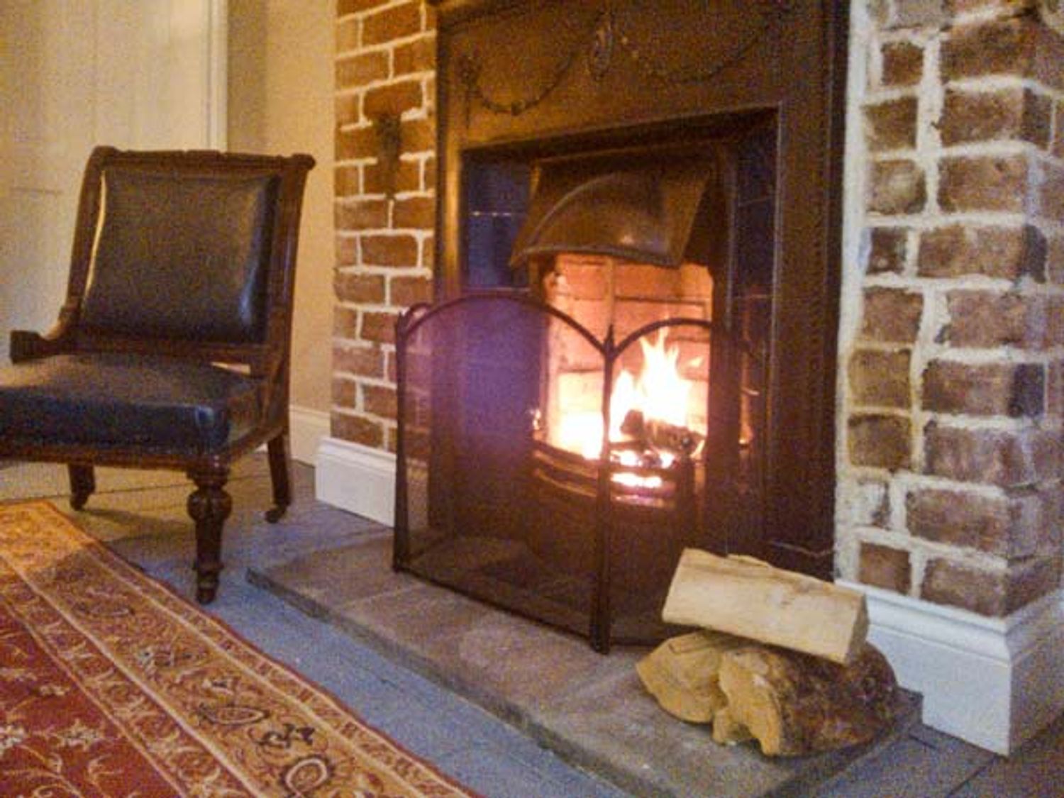 A fireplace with firewood and fireguard beside a chair on a carpet at Woodland Cottage in Malvern