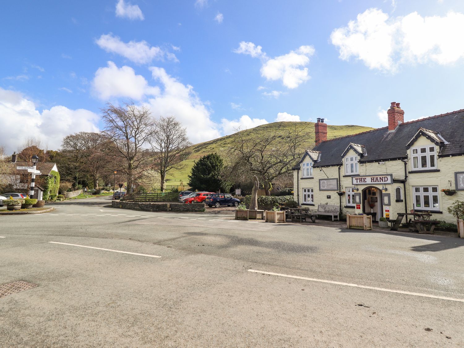 A street with parked cars trees and a building named The Hand near a hill at Minffordd in Llanarmon Dyffryn Ceiriog