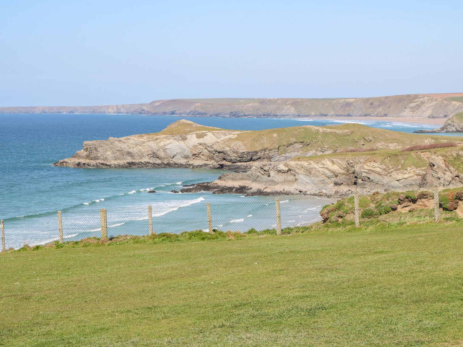 A coastal view with a grassy field and a wire fence overlooking rocky cliffs and the sea at The Beach House in Porth near Newquay