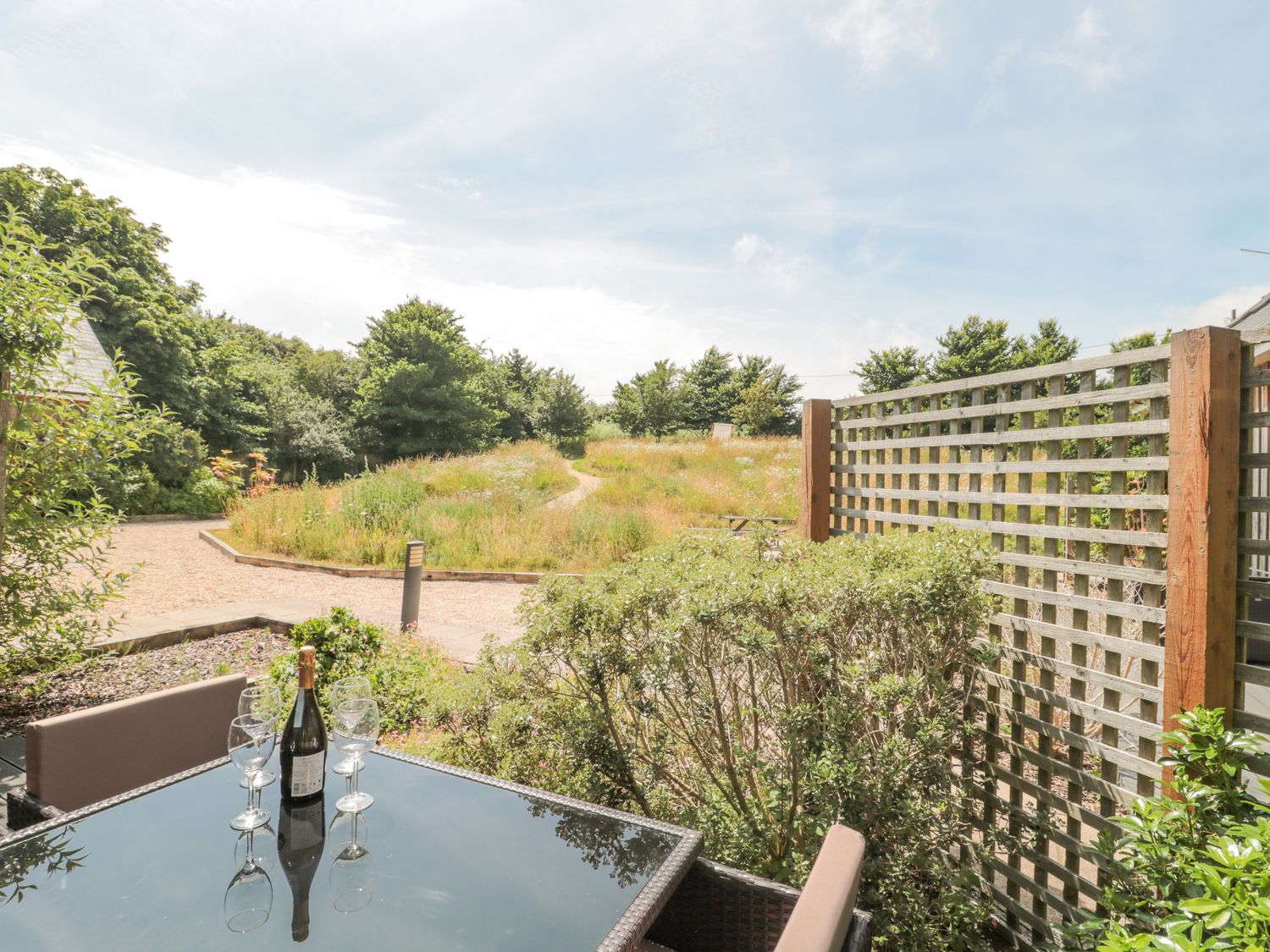 An outdoor patio with a glass table holding a bottle and wine glasses next to a lattice fence and greenery at Everdene Upwey near Weymouth
