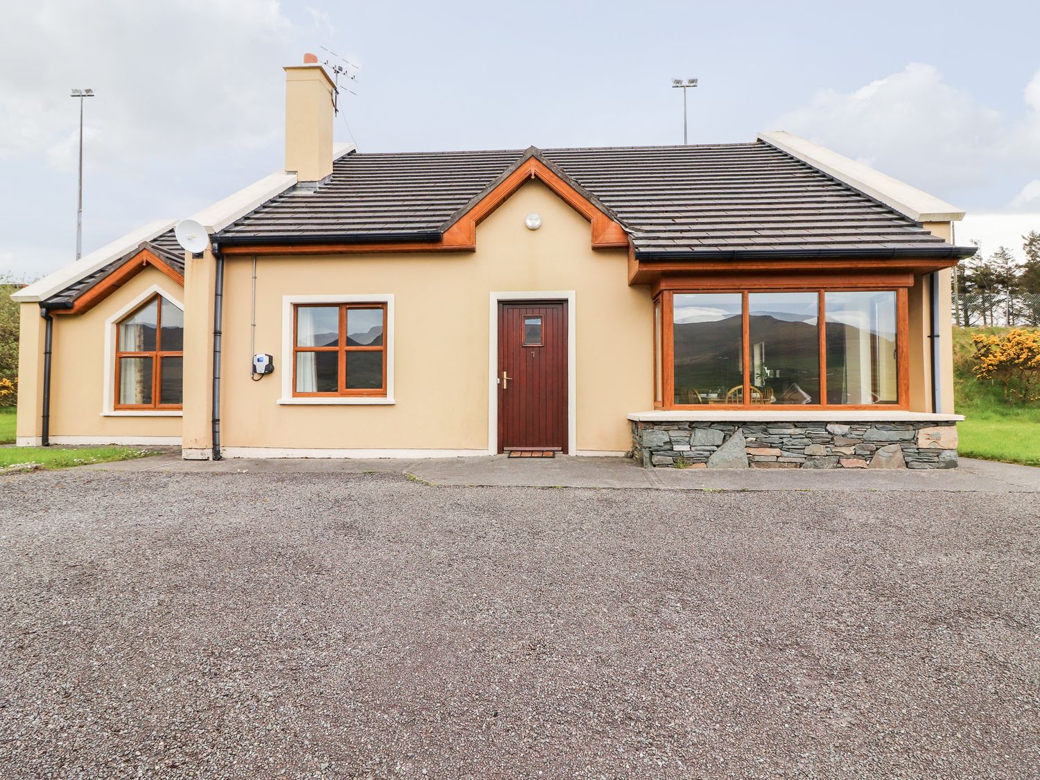 A single-story house with a brown front door and multiple windows surrounded by a gravel driveway at Number 7 in Lispole County Kerry