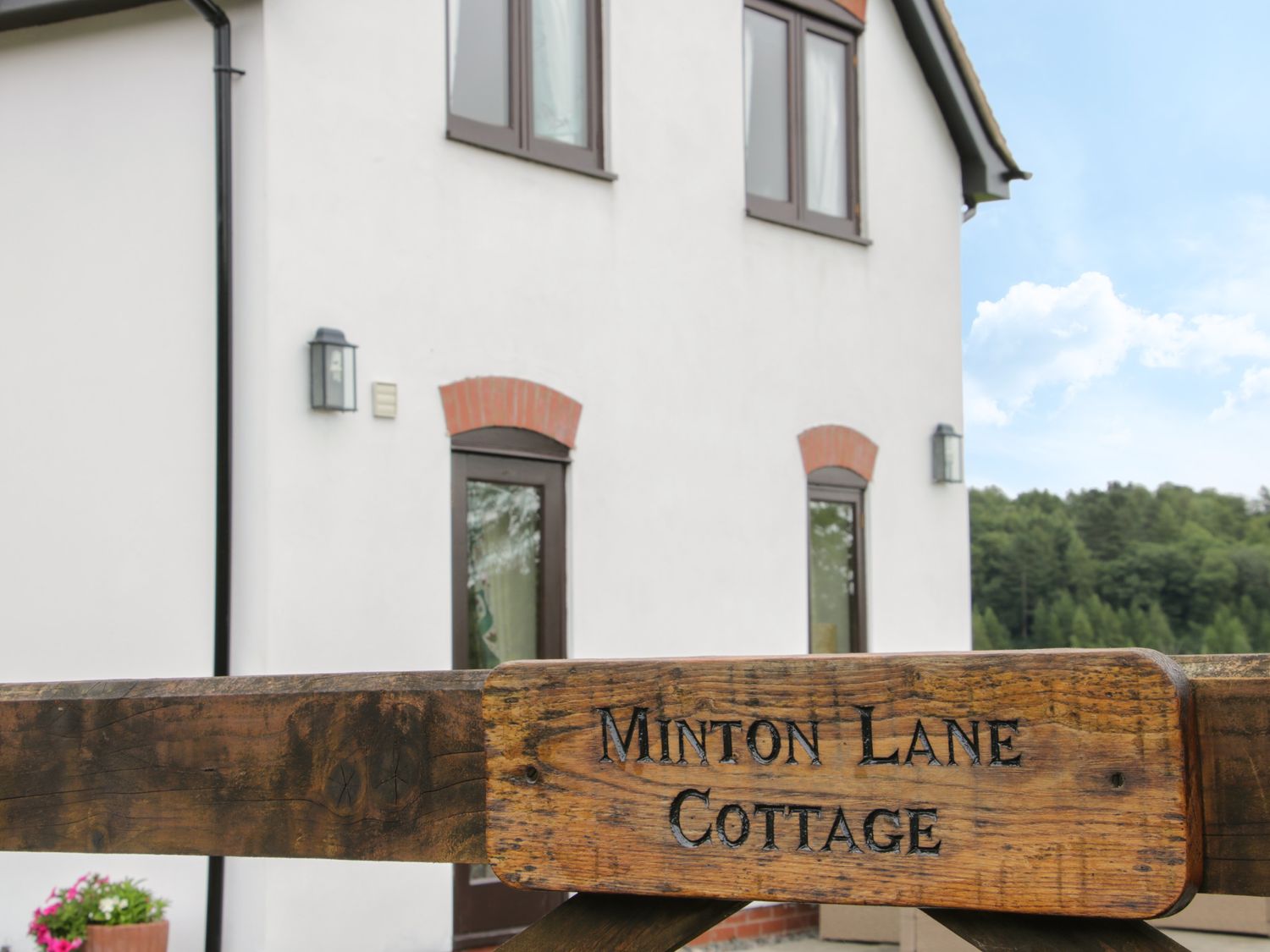 A wooden sign with Minton Lane Cottage in front of a white house with windows and outdoor lights at Minton Lane Cottage in Little Stretton
