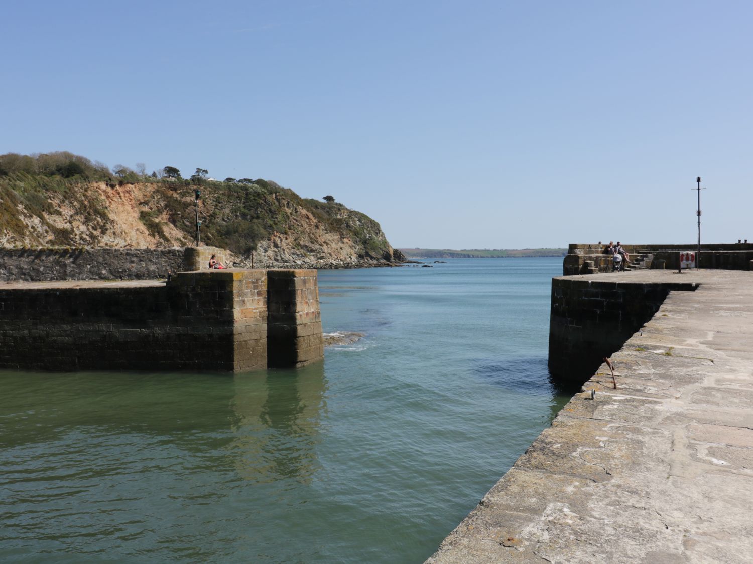 A harbor with stone piers and people sitting on the edges near water and cliffs at The Mackerel in Charlestown