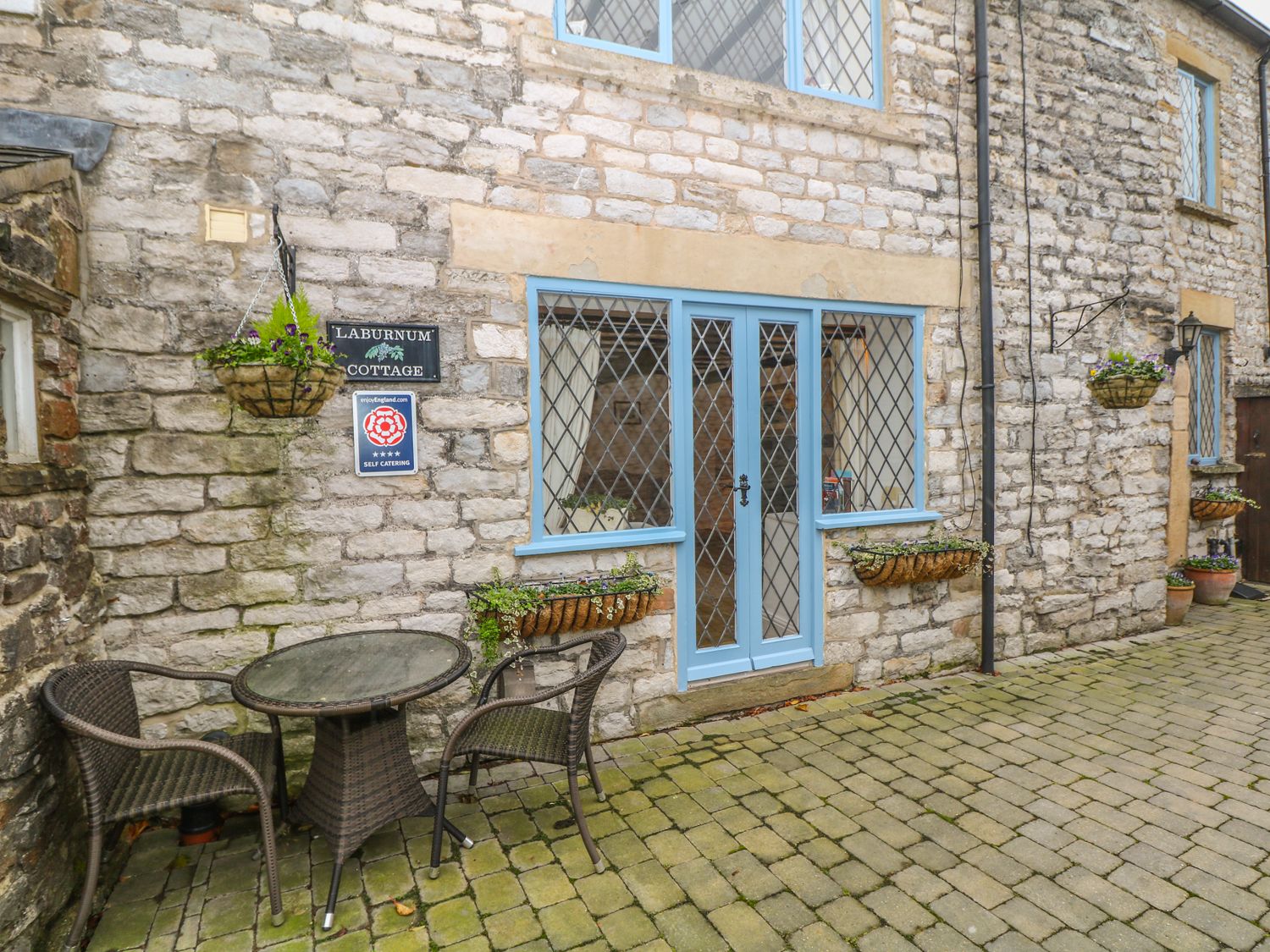 An outdoor stone patio with a glass table and two wicker chairs outside a stone cottage with blue framed windows and door at Laburnum Cottage in Castleton