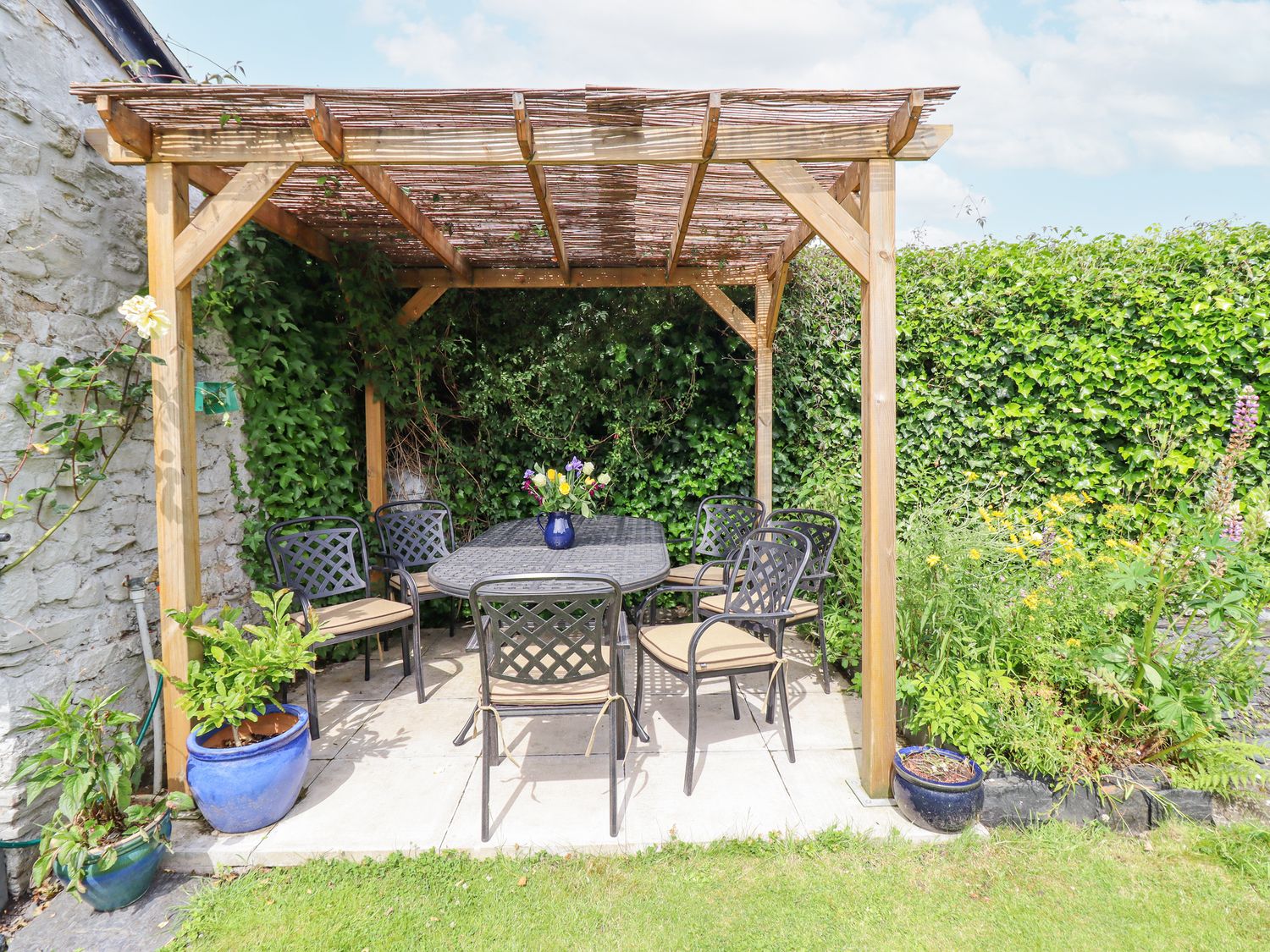 A wooden pergola with a metal table and six chairs on a tiled patio surrounded by potted plants and greenery at Bodywen in Llangynog