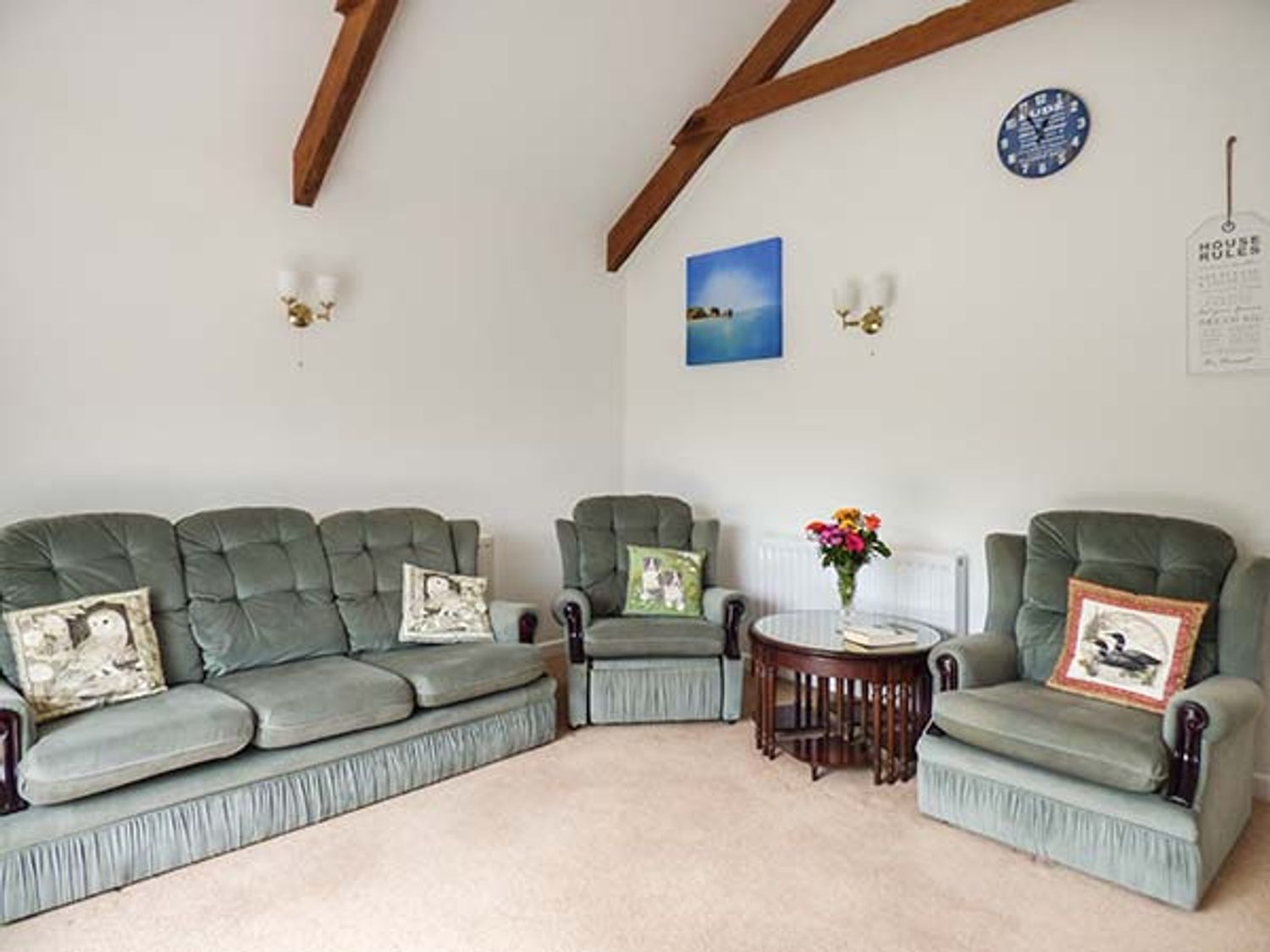 A living room with green upholstered sofa and chairs with pillows and a wooden round table with flowers at Shepherds Rest in Jacobstow near Bude