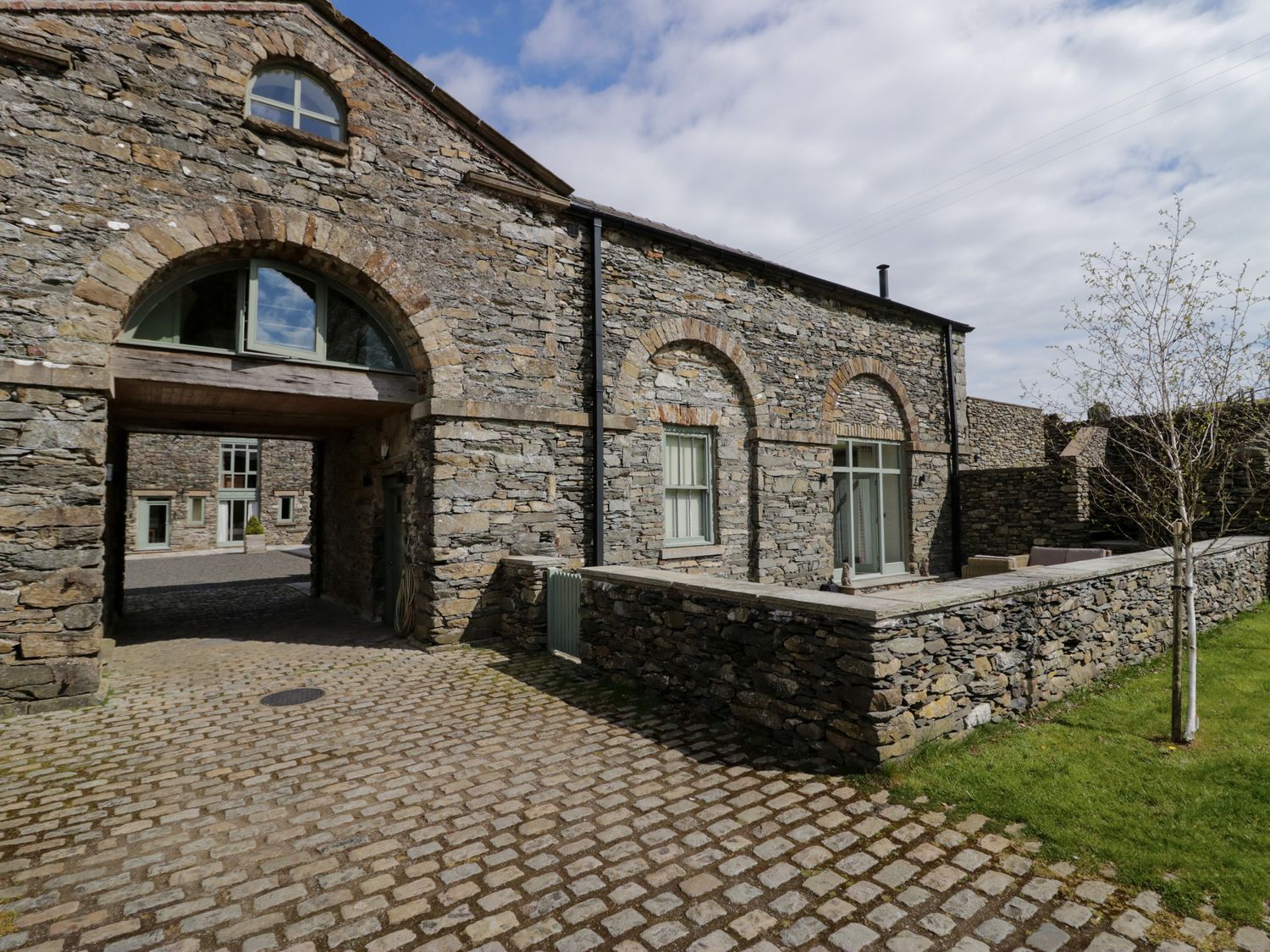 A stone building with arched windows and an archway over a cobblestone path at Rowan Cottage in Backbarrow near Newby Bridge